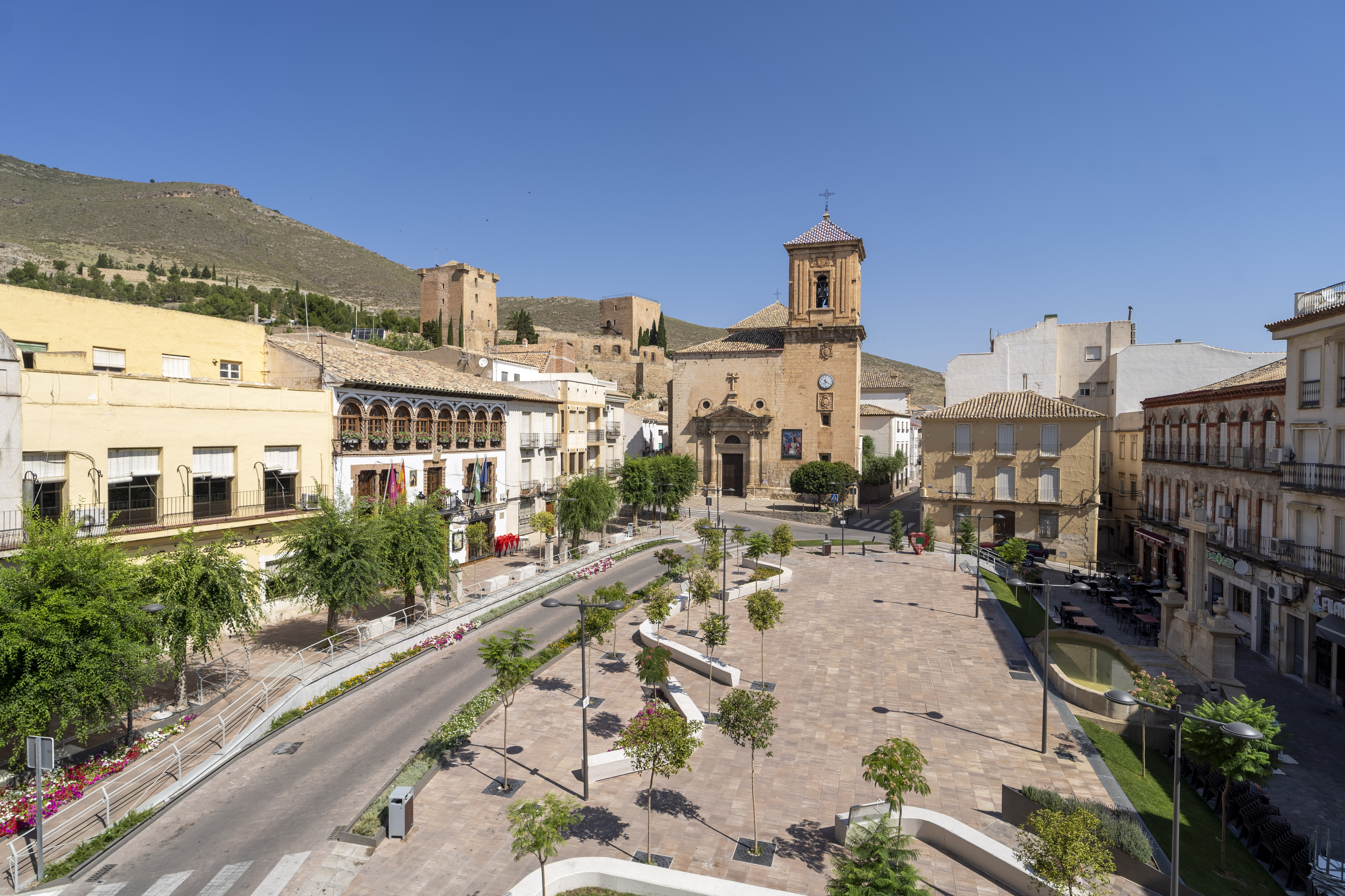 La imagen muestra una plaza urbana de carácter histórico, presidida por una iglesia y rodeada de edificios tradicionales perfectamente integrados en el paisaje. Al fondo destacan las antiguas torres y fortificaciones sobre la ladera, que aportan una fuerte identidad patrimonial y monumental al conjunto.