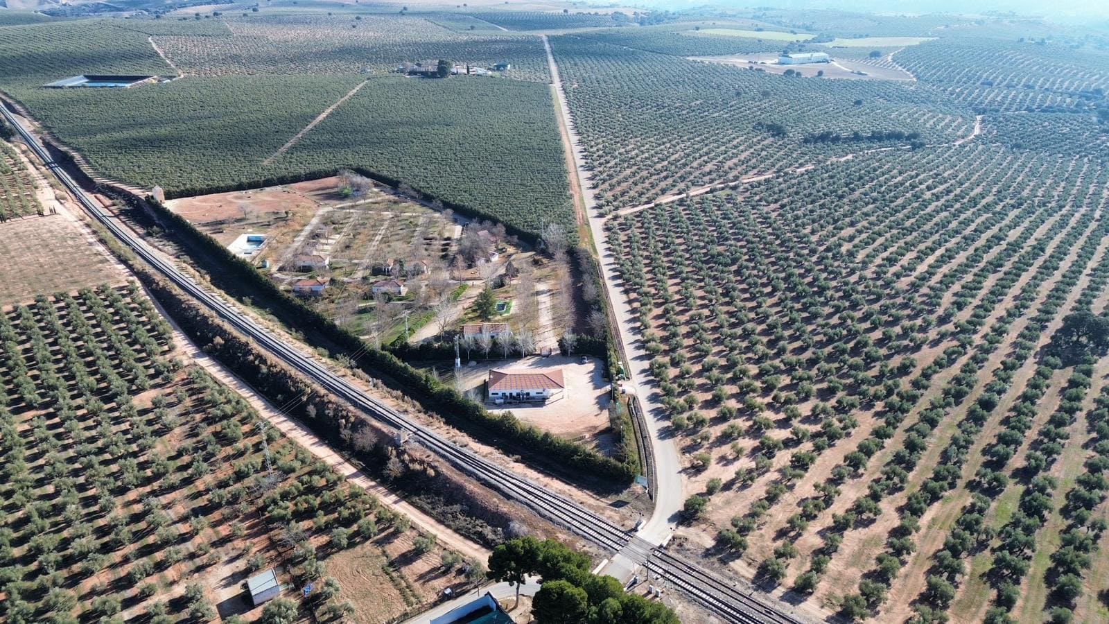 Vista aérea de una finca rodeada de extensos olivares, con un edificio principal junto a una carretera y una vía de tren que atraviesa el paisaje.