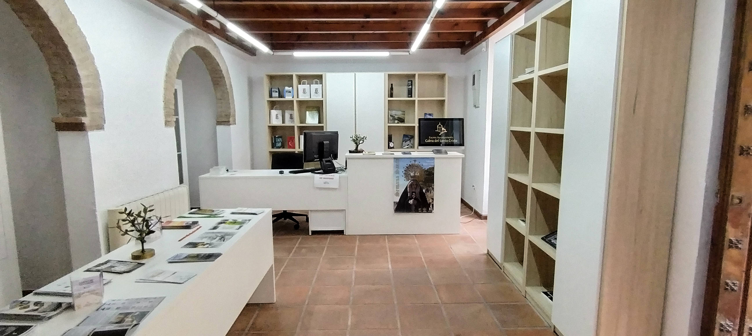 Interior de una oficina de turismo con un espacio cuidado y funcional, donde destacan los arcos de piedra, el techo de vigas de madera y la combinación de elementos tradicionales y modernos. El mostrador de atención y las estanterías organizan folletos e información para visitantes, creando un ambiente acogedor y ordenado orientado a la atención turística.