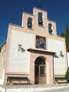 Ermita de pequeñas dimensiones y estética tradicional, con fachada blanca rematada por una espadaña de tres campanas. Sobre la entrada principal, enmarcada por piedra, destaca un mosaico cerámico de carácter religioso que aporta un elemento decorativo singular. El acceso se realiza a nivel de calle y está flanqueado por bancos, lo que sugiere un espacio de reunión tranquilo en el entorno inmediato.