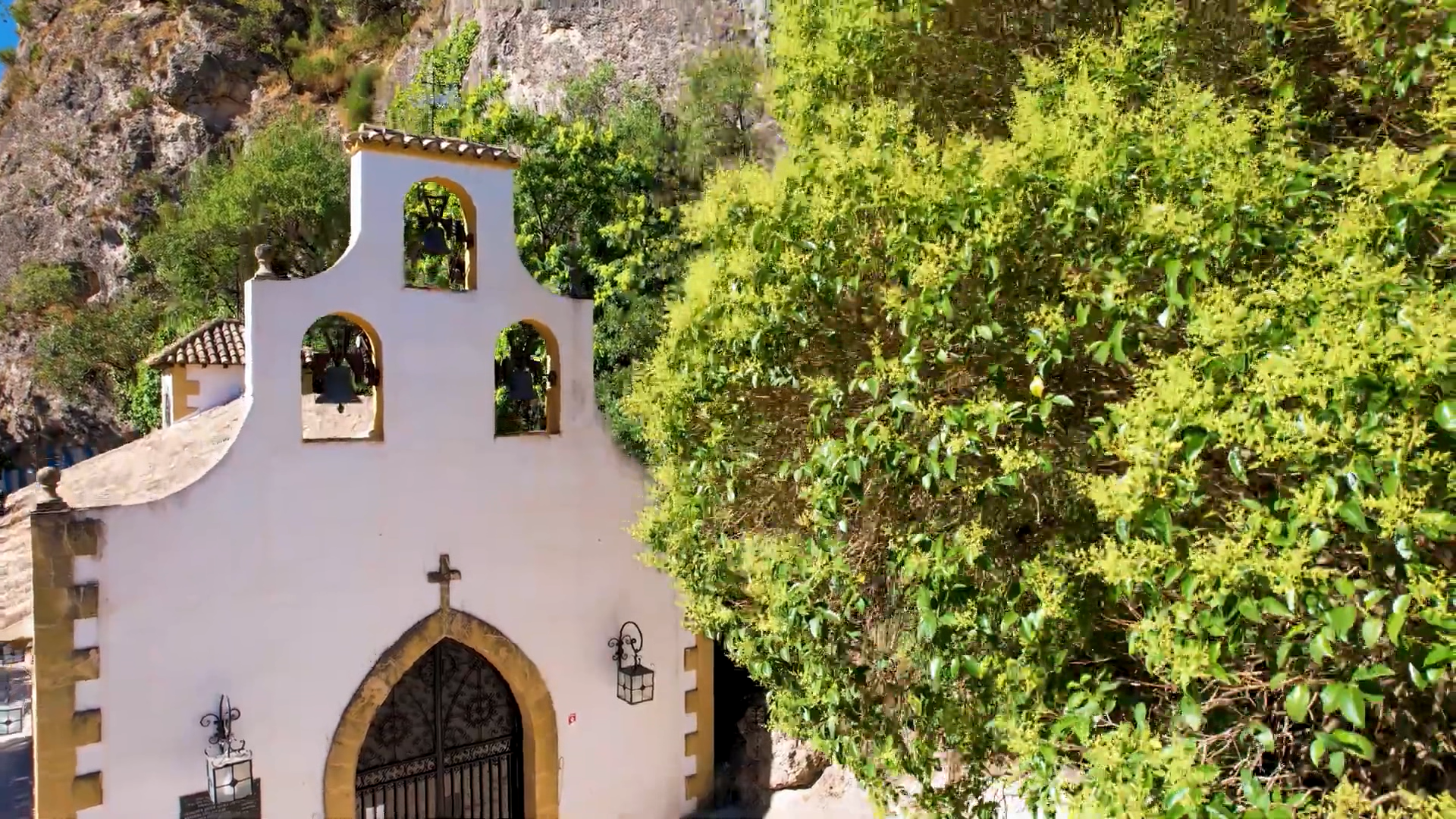 Pequeña ermita de fachada blanca y líneas sencillas, coronada por una espadaña con campanas, que se alza integrada en la ladera rocosa. La entrada, con arco de piedra y cruz superior, refuerza su carácter tradicional, mientras la vegetación que la rodea aporta frescura y resalta su armonía con el entorno natural de la sierra.