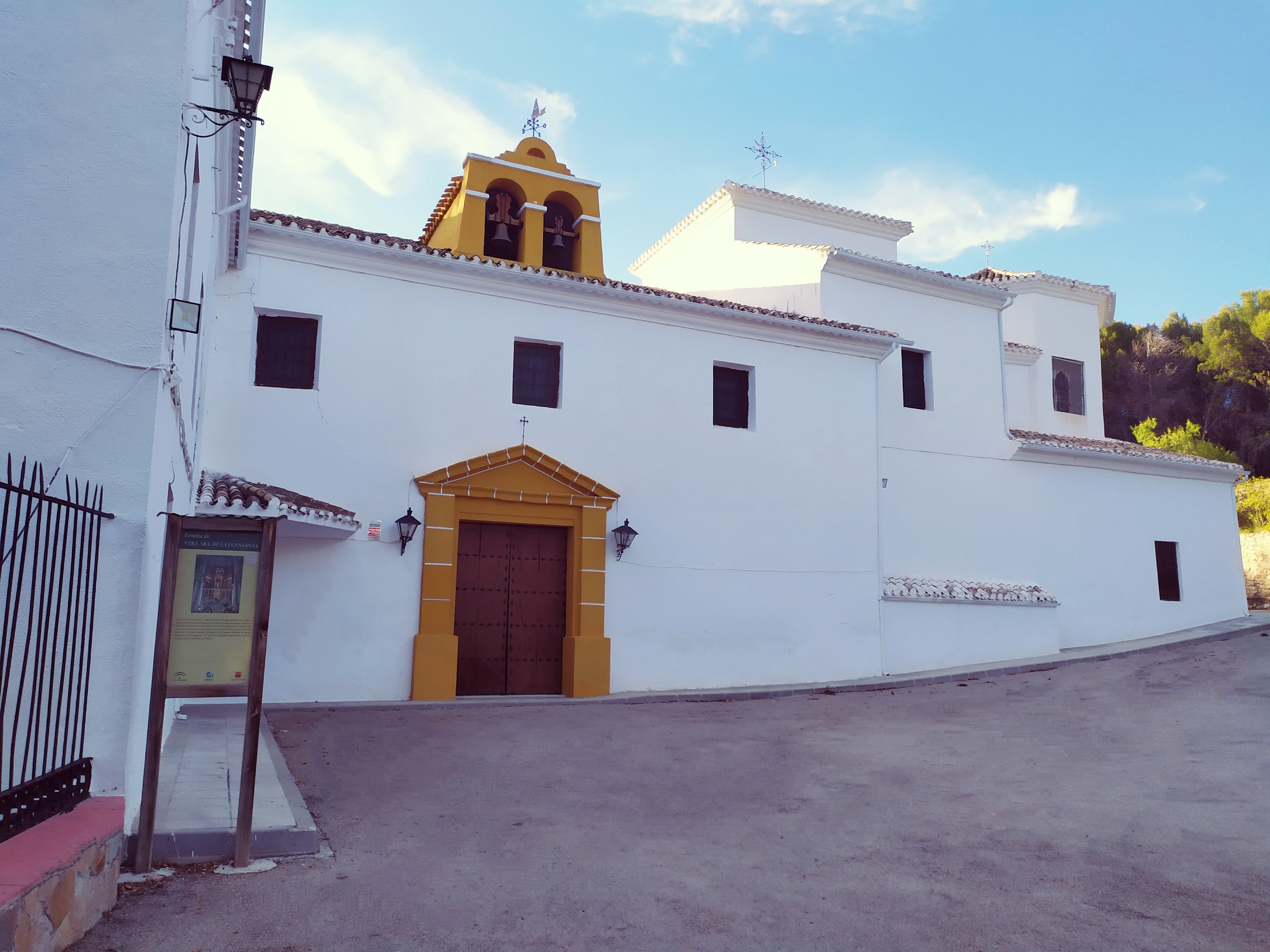 Fachada de la Ermita de Nuestra Señora de la Fuensanta, un edificio de paredes encaladas situado en un entorno tranquilo y natural. Destaca la puerta principal enmarcada en tonos ocres y la pequeña espadaña con campanas que corona el templo, rodeado de vegetación propia de Sierra Mágina.