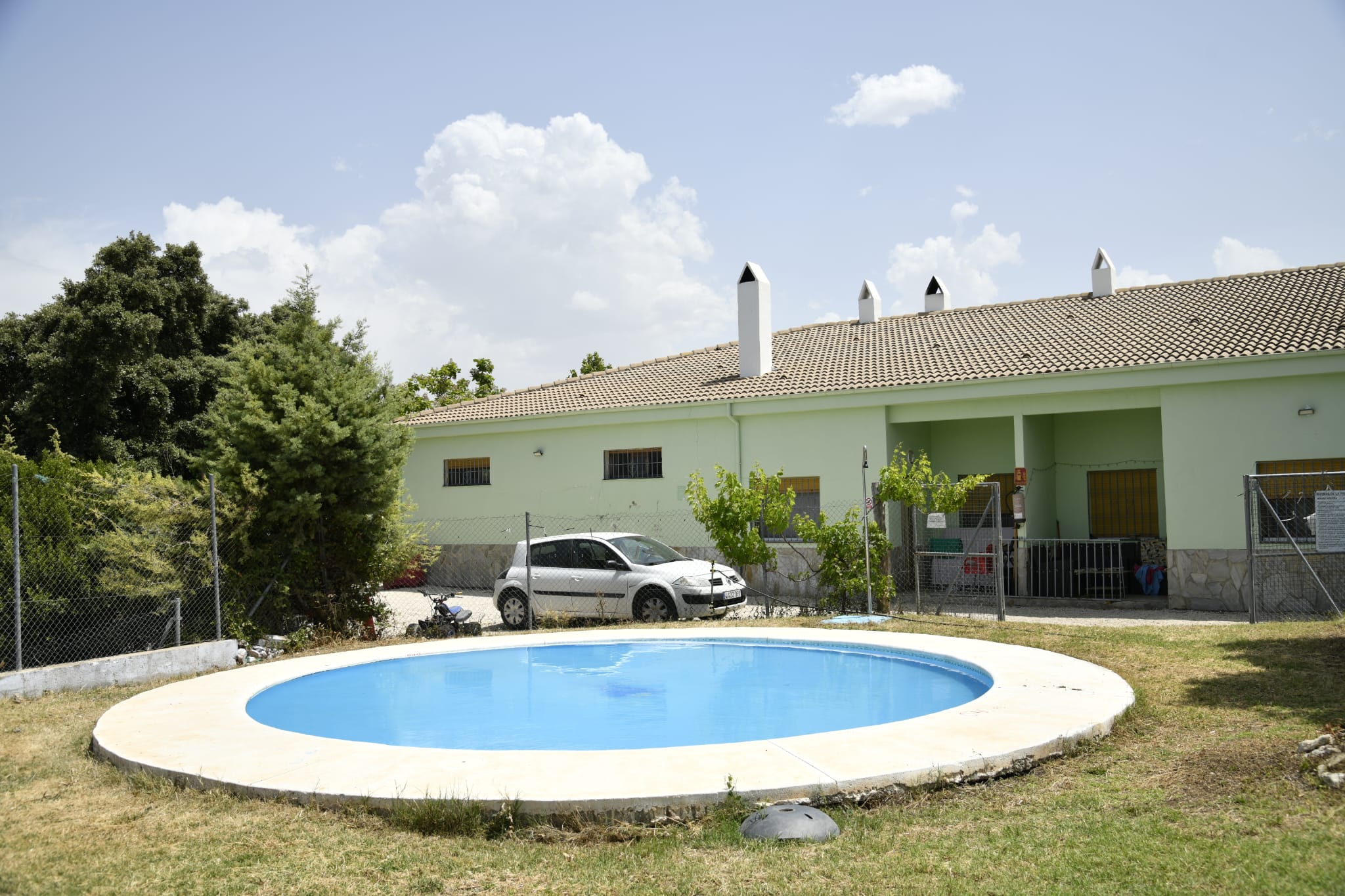 Zona de piscina circular en el camping, situada frente a un edificio de una sola planta con estética sencilla y funcional. El espacio está rodeado de vegetación y áreas de sombra, creando un ambiente tranquilo y familiar, ideal para el descanso tras una jornada de actividad en la sierra.