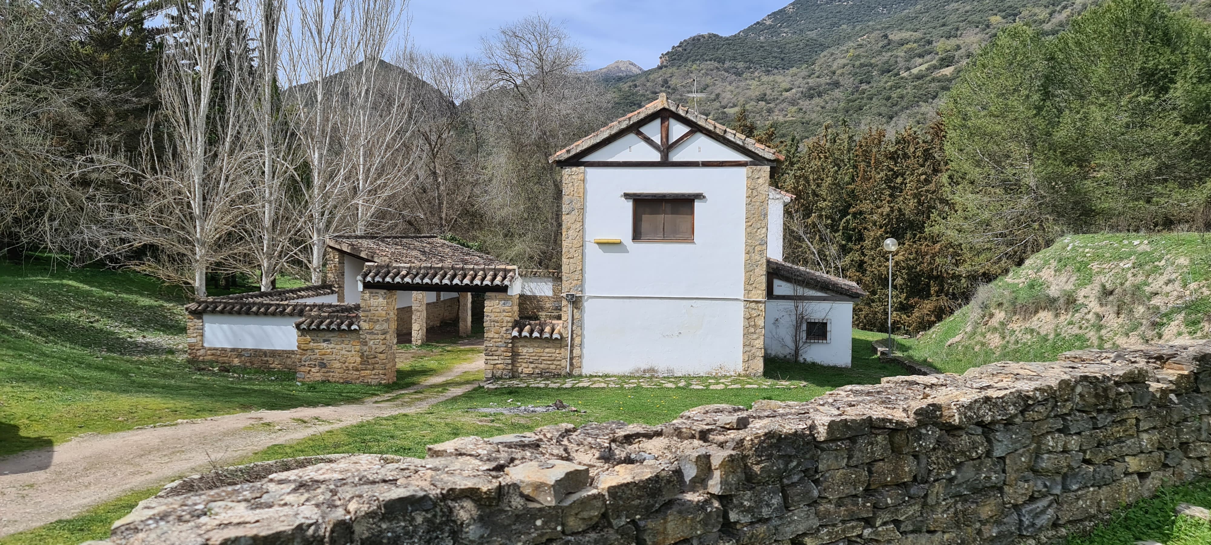 Edificación tradicional de piedra y fachada blanca situada en un entorno natural, rodeada de prados y arbolado al pie de la sierra. El conjunto incluye un pequeño porche con cubierta de teja y muros de mampostería, evocando la arquitectura rural ligada a antiguos usos agrícolas y ganaderos, en un paisaje tranquilo y abierto.