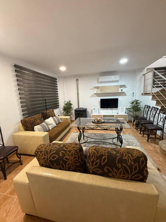 Salón amplio y luminoso de estilo actual, con sofás en tonos beige y estampados, mesa de centro de cristal y zona de televisión. El espacio combina elementos modernos con detalles funcionales, como aire acondicionado y estufa, creando un ambiente cómodo y preparado para el descanso en grupo.