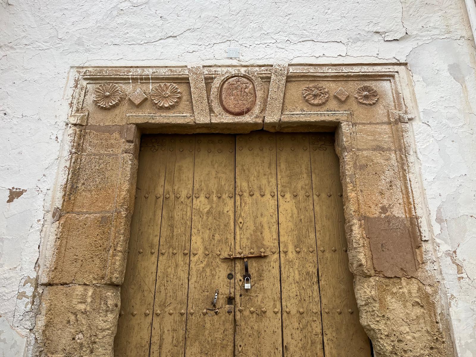Detalle de la portada principal en piedra, con un dintel decorado mediante relieves geométricos y motivos florales. En la parte central aparece un medallón con inscripción religiosa, mientras que la puerta de madera conserva herrajes tradicionales y elementos metálicos originales. La textura de la piedra y el desgaste visible aportan un notable valor histórico y artesanal al conjunto.