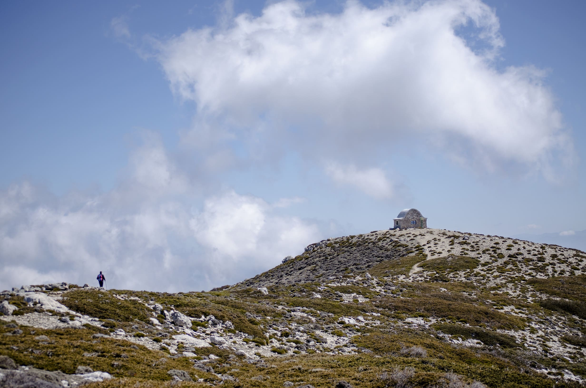 Paisaje de alta montaña en Sierra Mágina, donde un senderista avanza por la ladera rocosa camino de la cumbre. En lo alto se distingue un pequeño refugio de piedra en el Pico Miramundos, rodeado de vegetación baja y bajo un cielo abierto con grandes nubes.