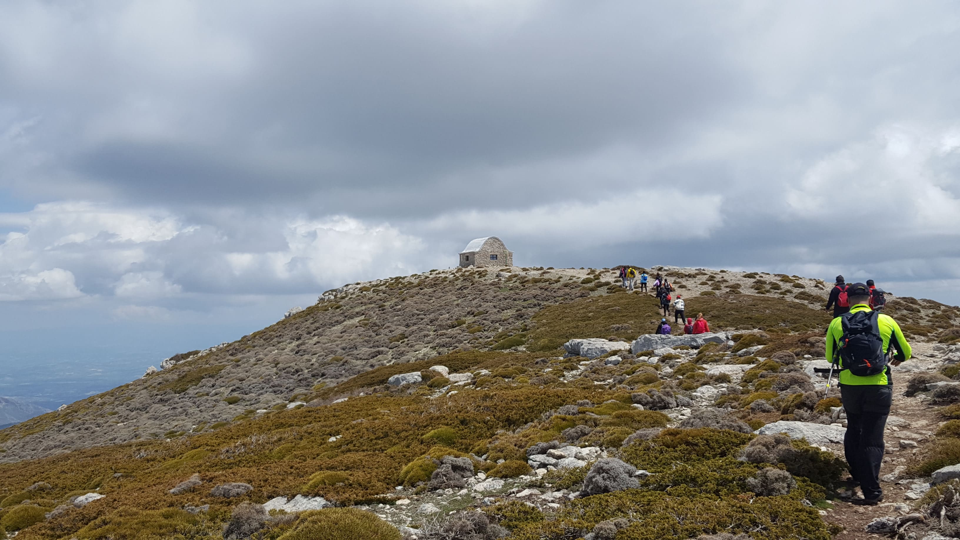rupo de senderistas recorriendo un sendero en cresta hacia una pequeña edificación en la cima, rodeados de amplias vistas del paisaje.
