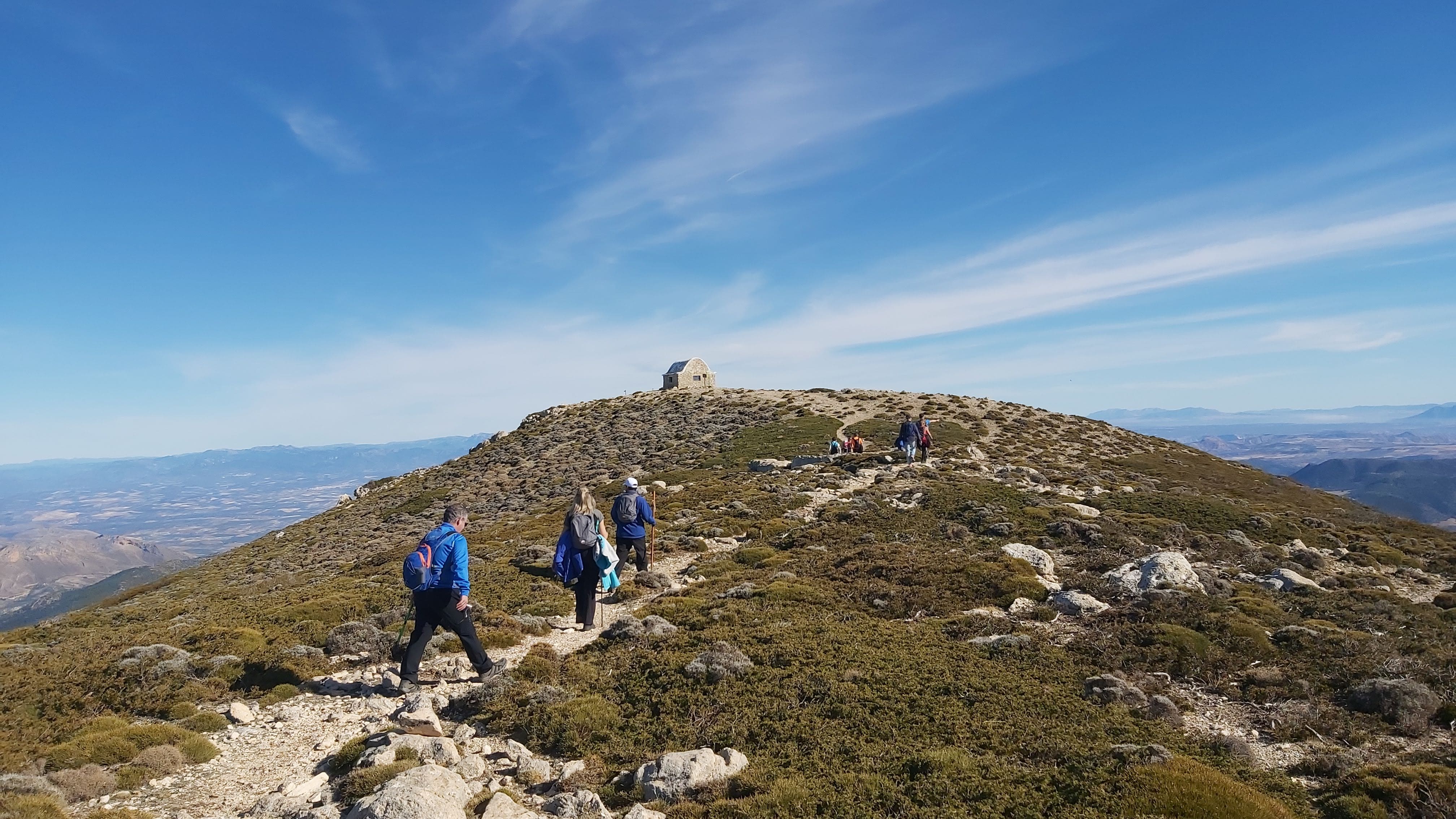 Varias personas caminan por un terreno pedregoso y abierto hacia una pequeña construcción en la cima, bajo un cielo cubierto que acentúa la sensación de altura.