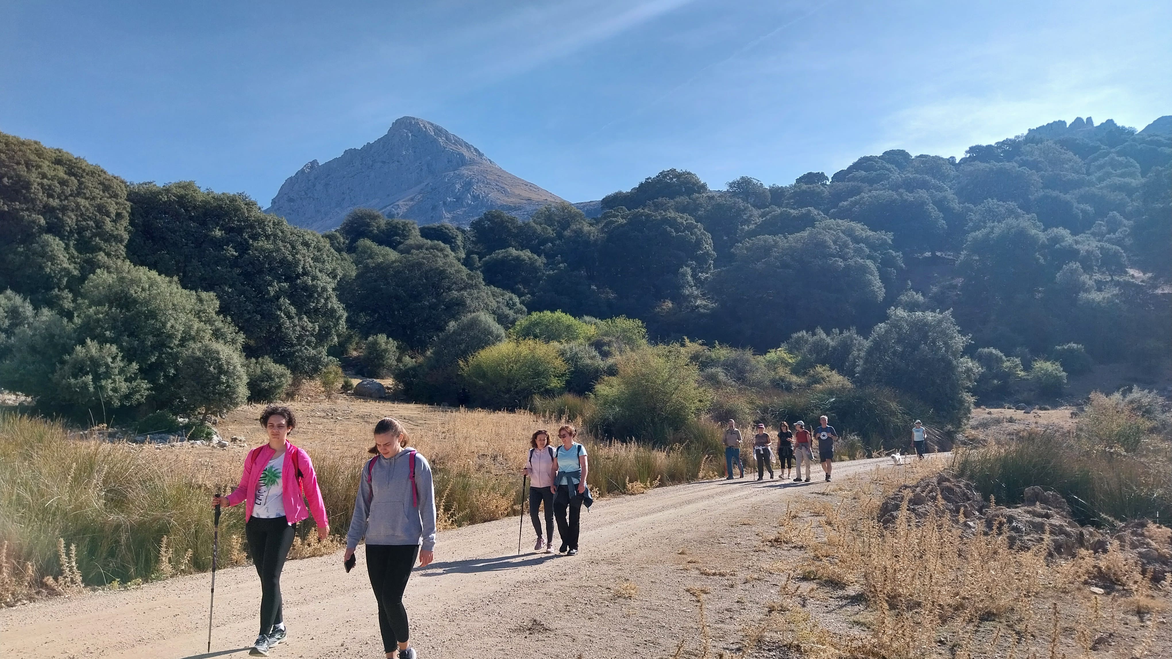 Senderistas recorren un camino amplio en un paisaje abierto, con una montaña destacada al fondo y vegetación mediterránea a ambos lados.
