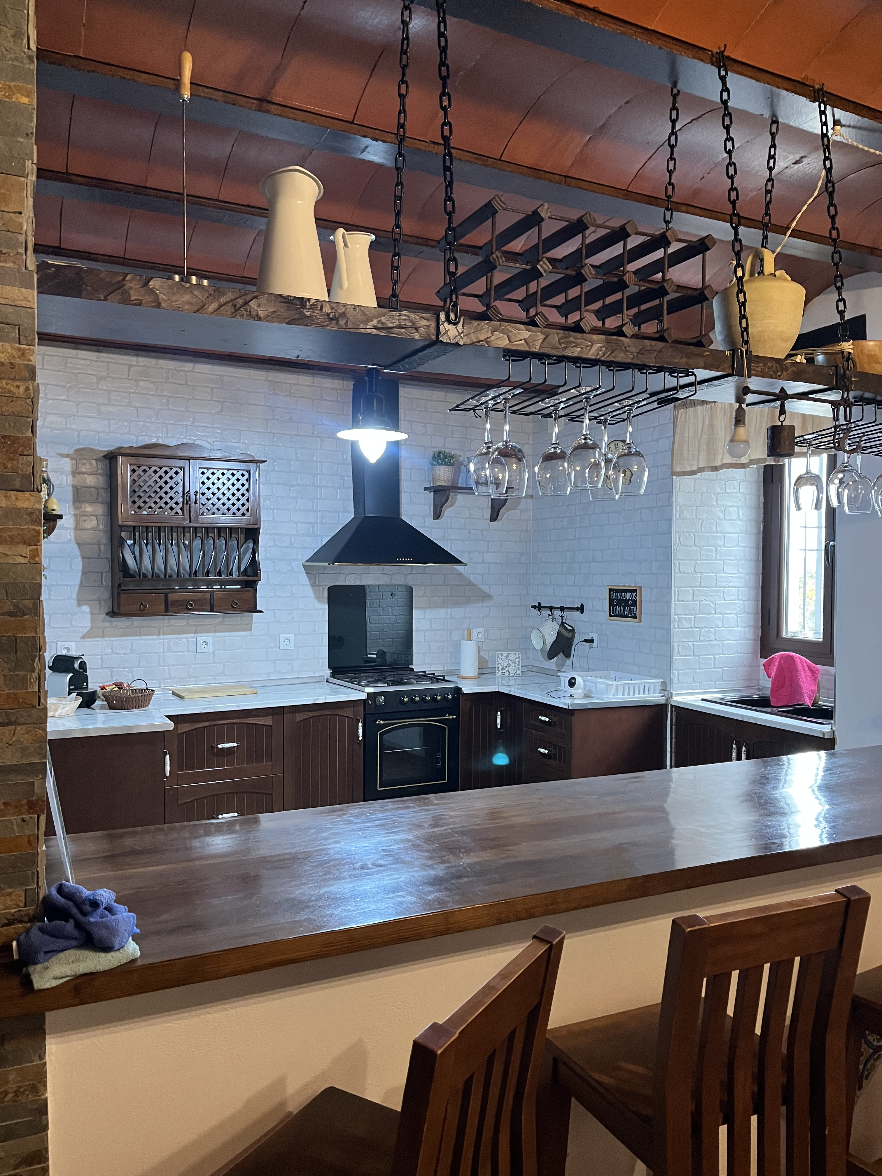 Vista de una cocina rústica y moderna, con una barra de madera, estanterías colgantes y una campana extractora negra. Hay copas y utensilios de cocina colgados sobre la barra, y el espacio está bien iluminado.