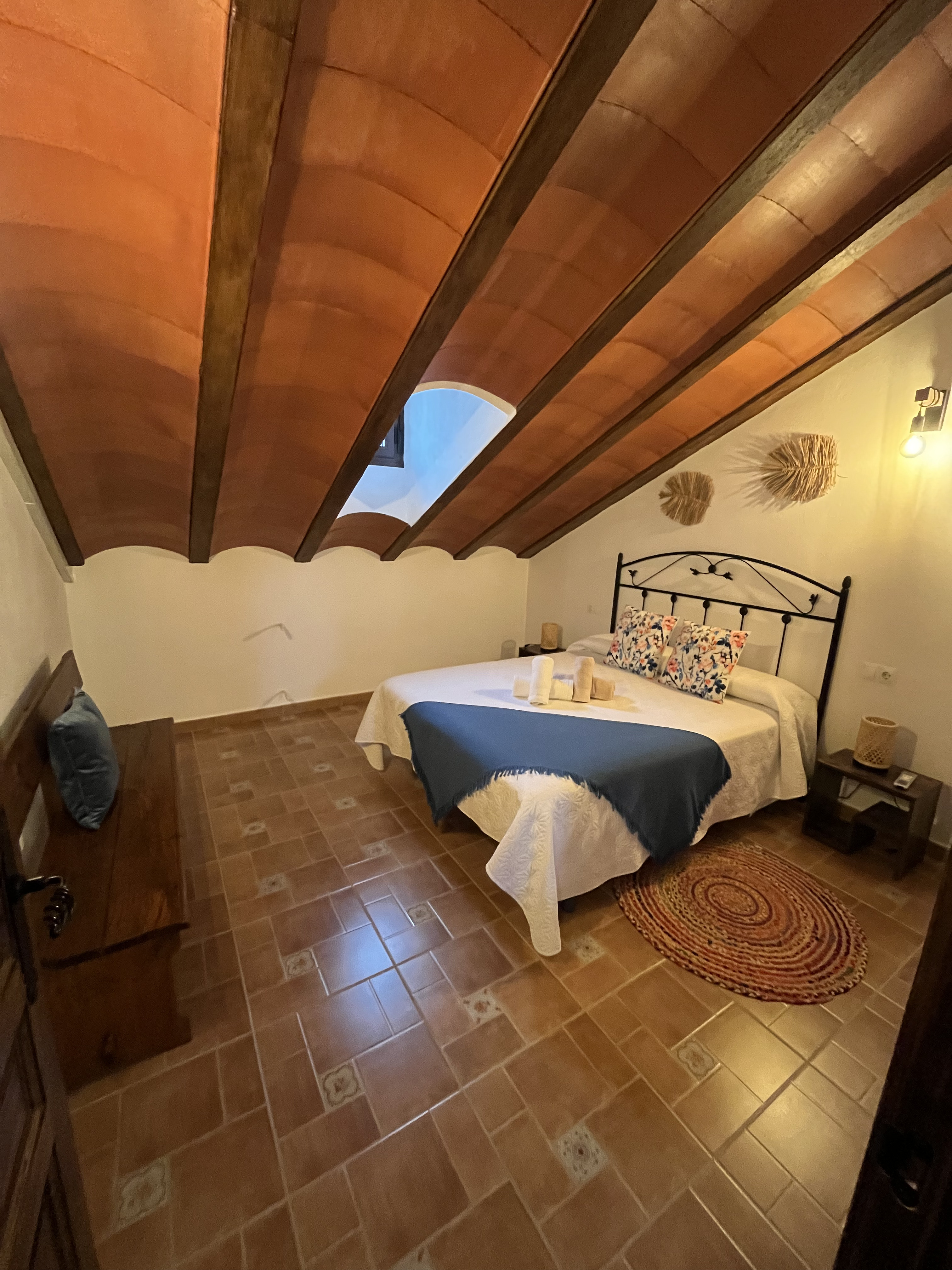 Otro dormitorio con techo abovedado y detalles de decoración sencilla. La cama tiene sábanas blancas y una manta azul, complementada con toallas dobladas en la cama. Hay una alfombra redonda en el suelo.