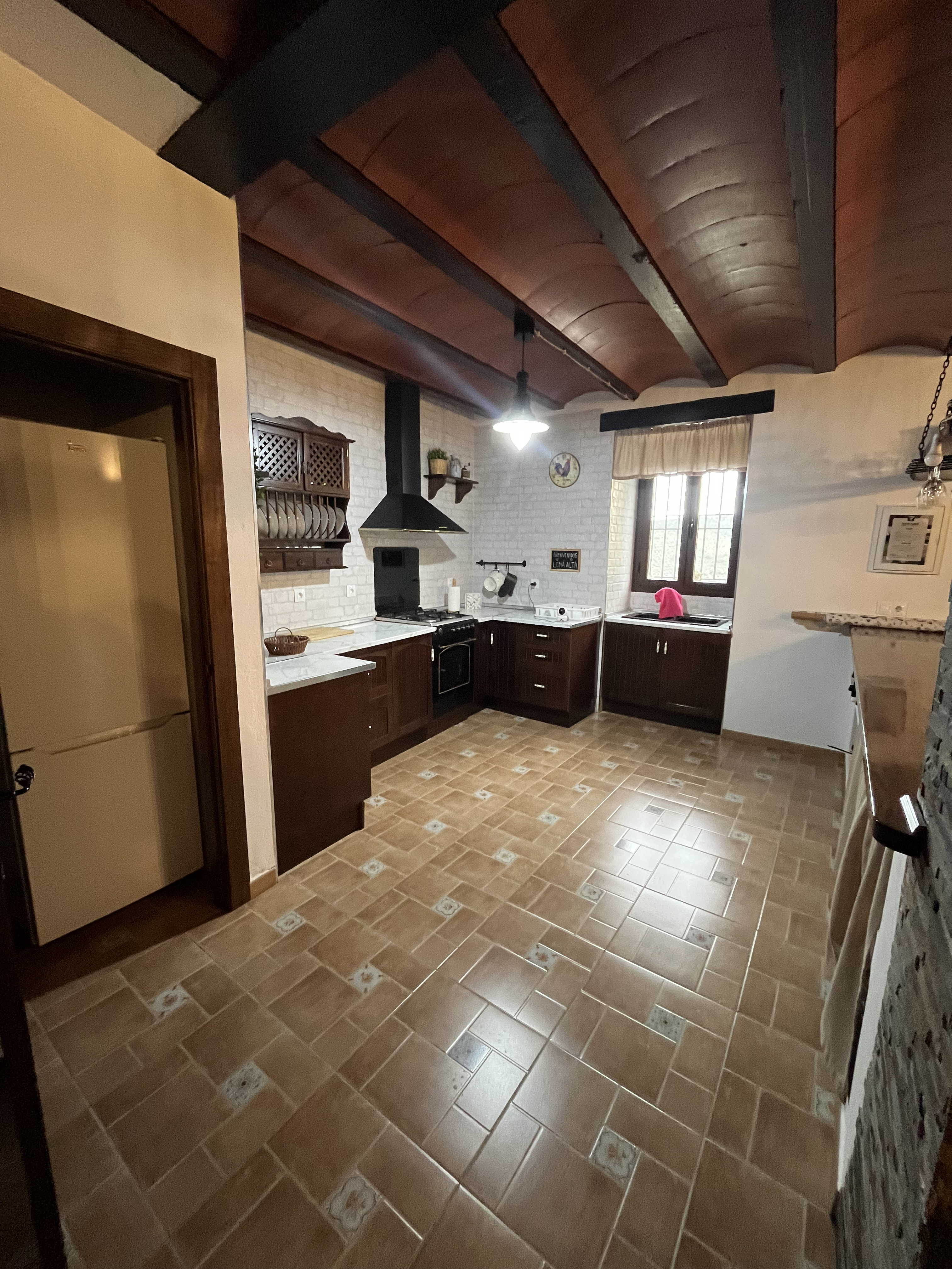 Cocina de estilo rústico con azulejos claros y madera oscura. Hay una isla central de madera, electrodomésticos modernos, y un ambiente acogedor, con ventana al exterior.