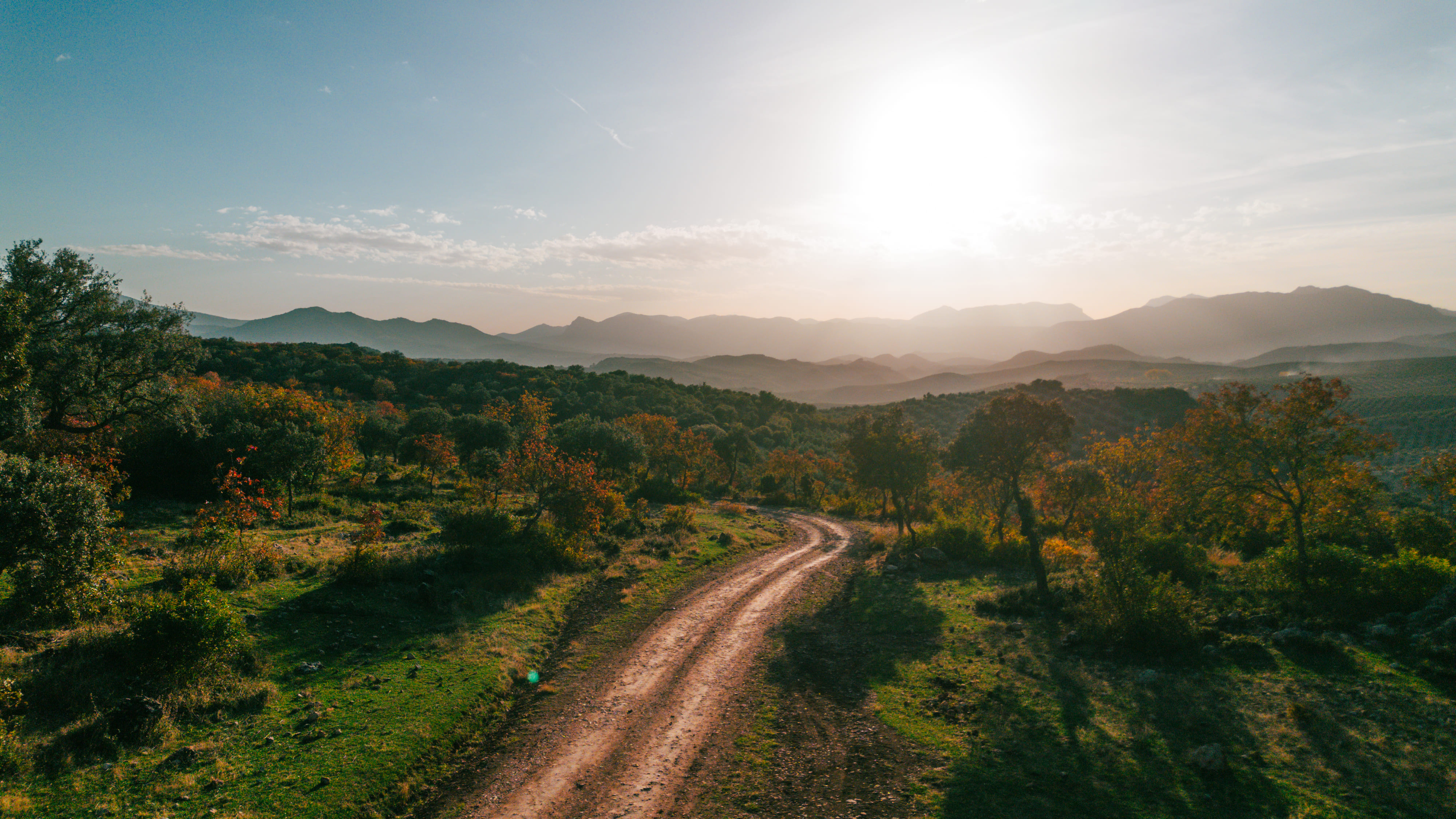 Camino de tierra que atraviesa un paisaje de vegetación y suaves lomas, abierto hacia un horizonte de sierras difuminadas por la luz. El sol alto y la atmósfera cálida aportan profundidad y una sensación de amplitud y tranquilidad.