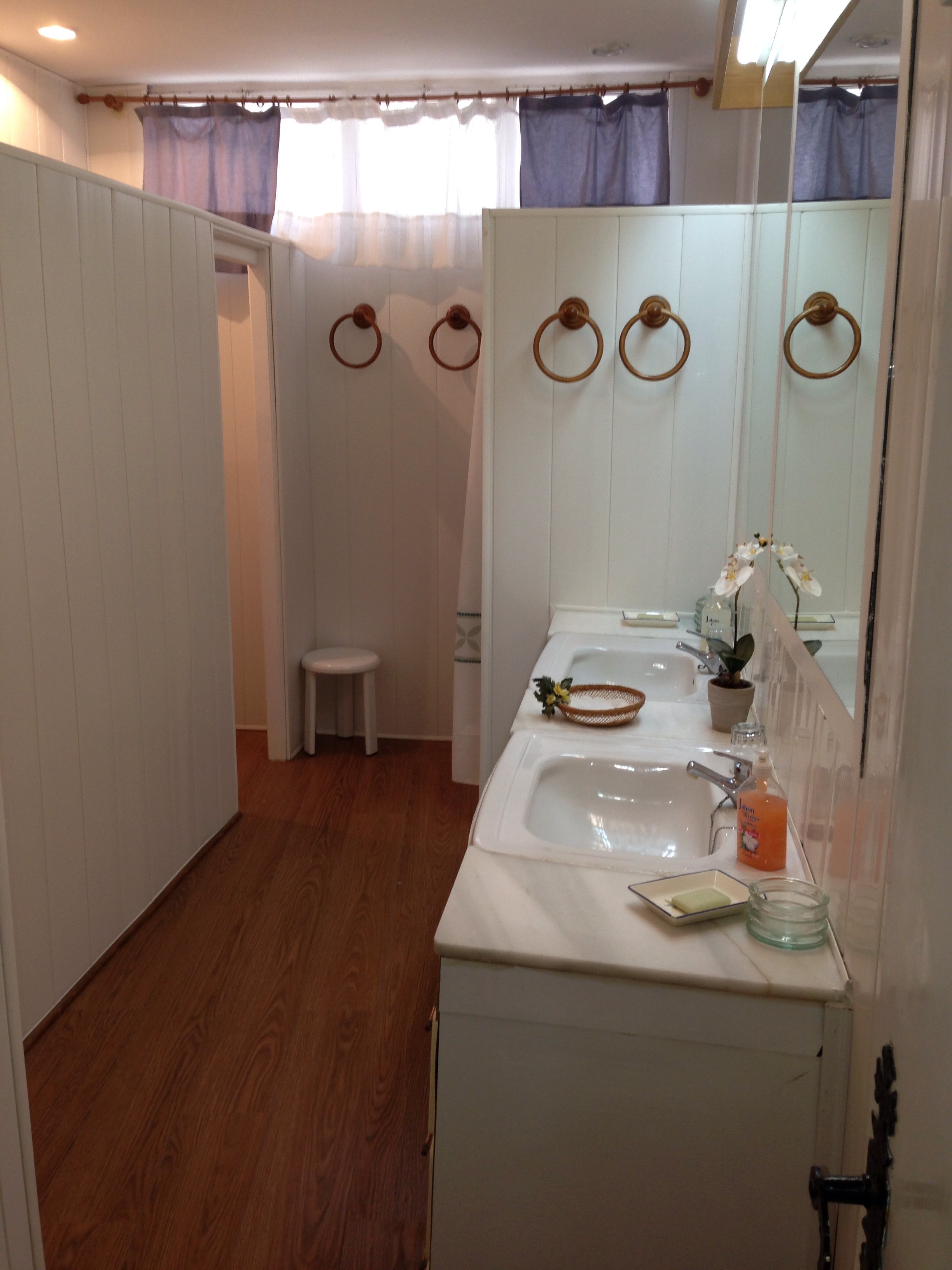 Espacio de baño amplio y luminoso, con doble lavabo sobre encimera de mármol y espejo corrido que amplía la sensación de profundidad. Predominan los tonos claros y la madera en el suelo, creando un ambiente cálido y funcional. Los detalles decorativos, como la planta y los accesorios, aportan un toque cuidado y acogedor.