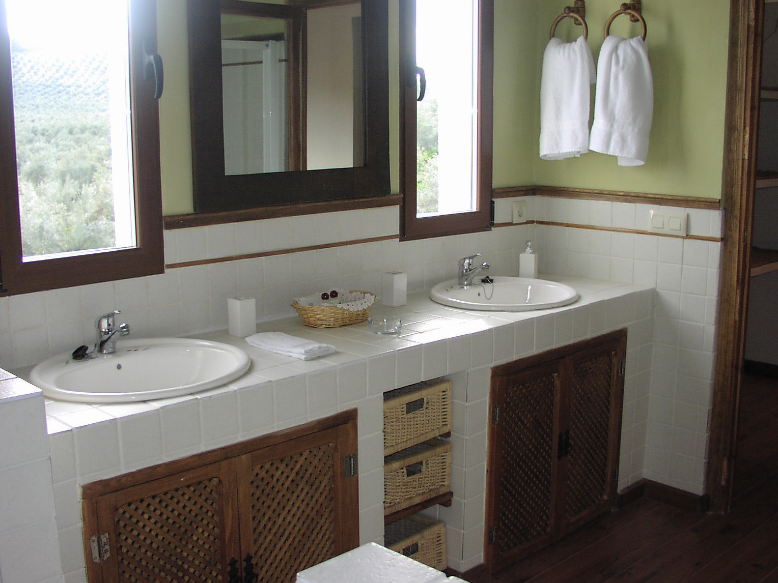 Baño amplio y luminoso con doble lavabo integrado en encimera de azulejo, acompañado de espejo de gran formato y detalles en madera. La luz natural entra por las ventanas, reforzando la sensación de limpieza y amplitud. Los elementos decorativos, como cestas y textiles, aportan un toque cálido y funcional.