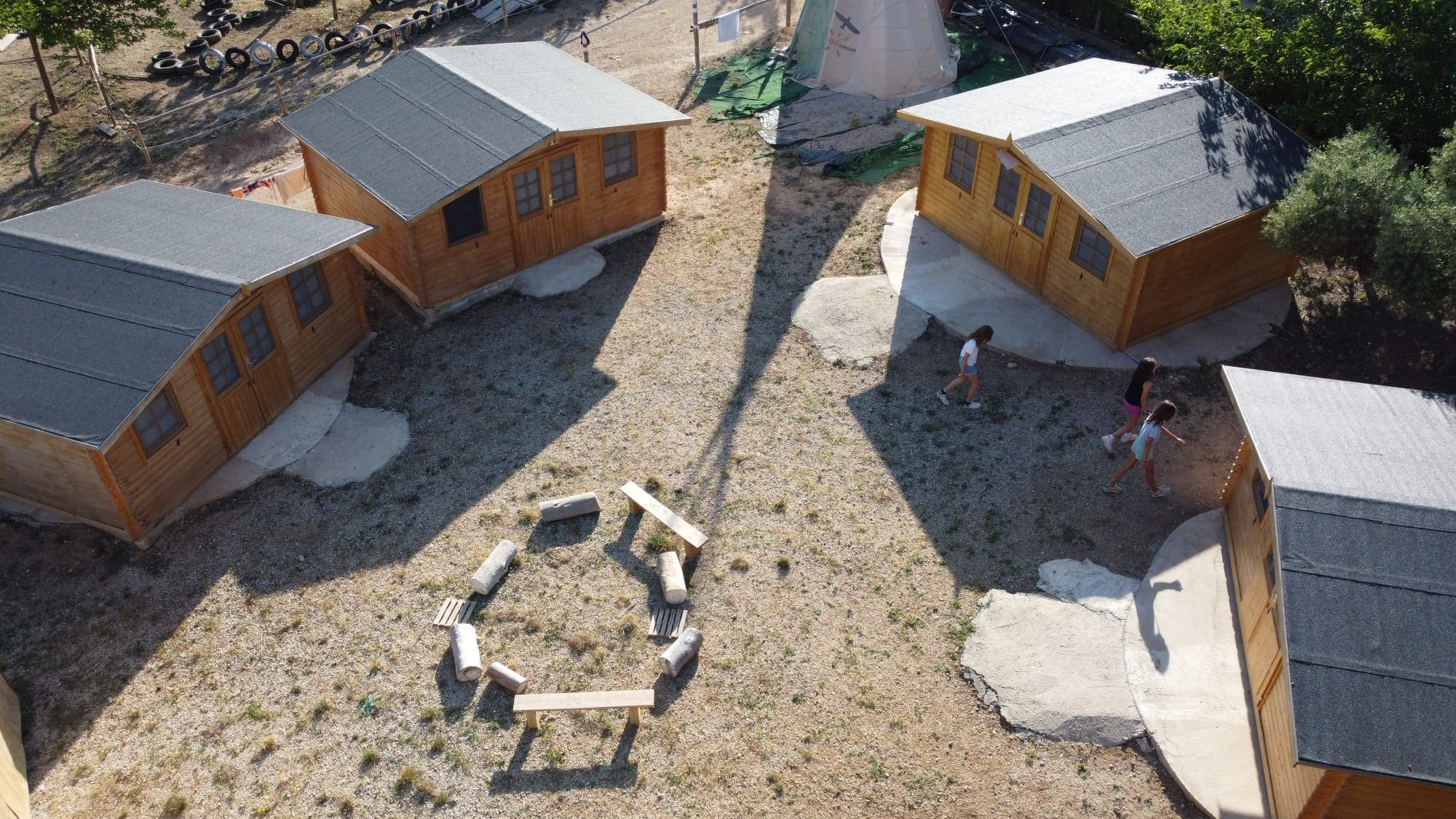 Espacio de alojamiento con pequeñas cabañas de madera distribuidas alrededor de una zona común con bancos, donde varios niños caminan entre ellas.