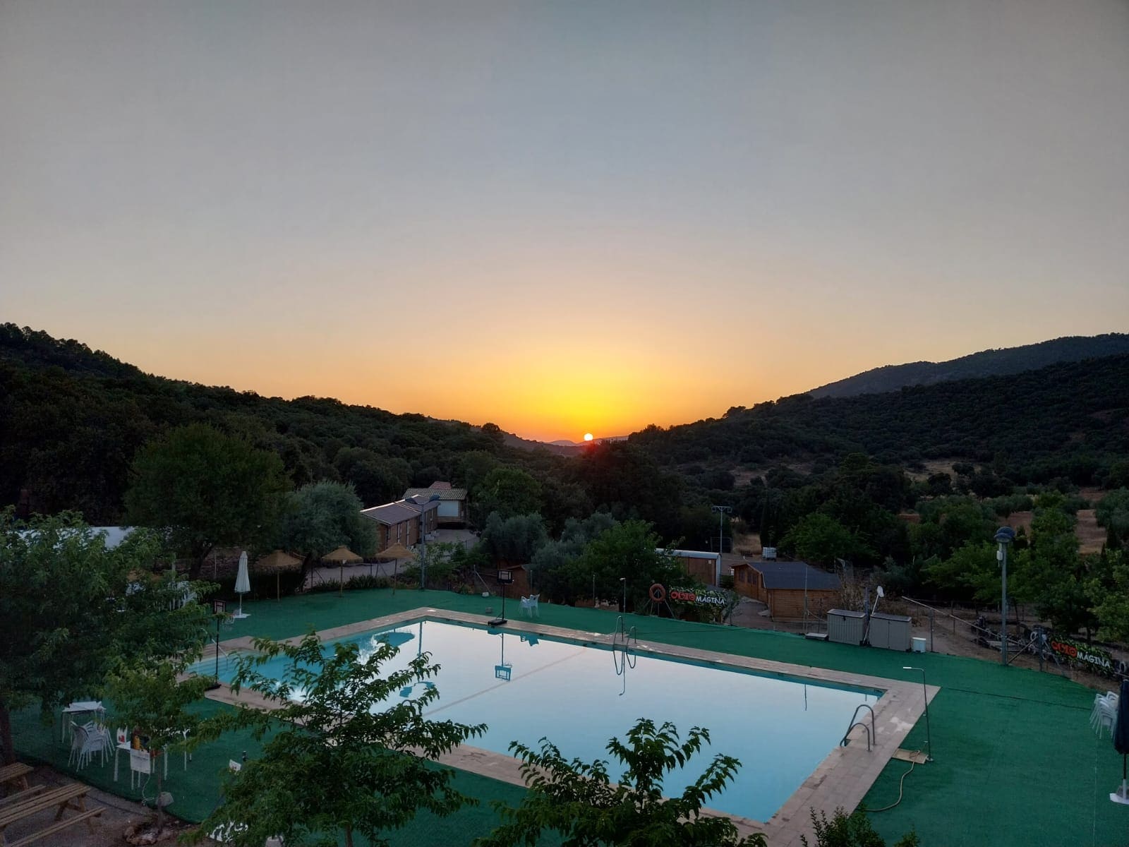 Piscina al aire libre al atardecer, con el sol ocultándose entre las montañas y un entorno natural de árboles y colinas.