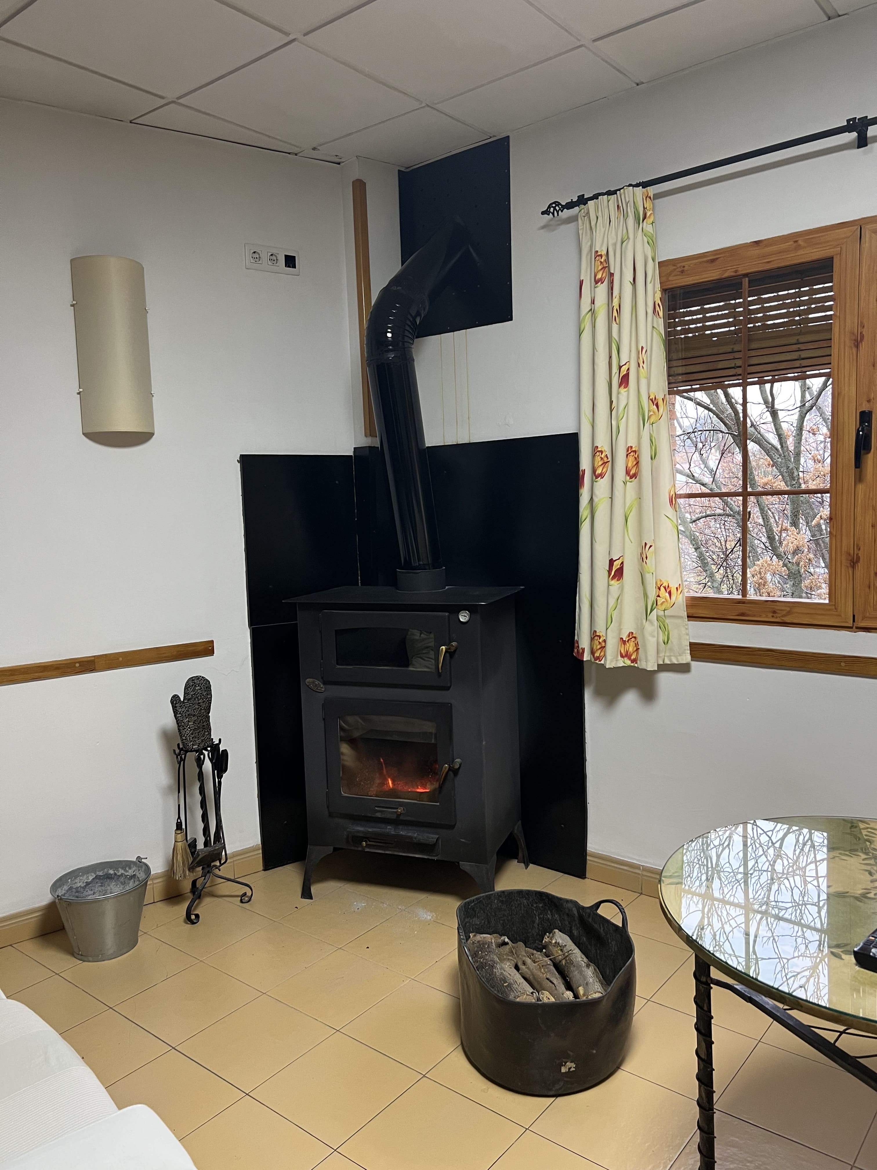 Una estufa de hierro negro está encendida, con una chimenea que se extiende hacia el techo. A su lado, hay un recipiente con madera y un cubo para el agua, mientras que la ventana muestra un paisaje con árboles.