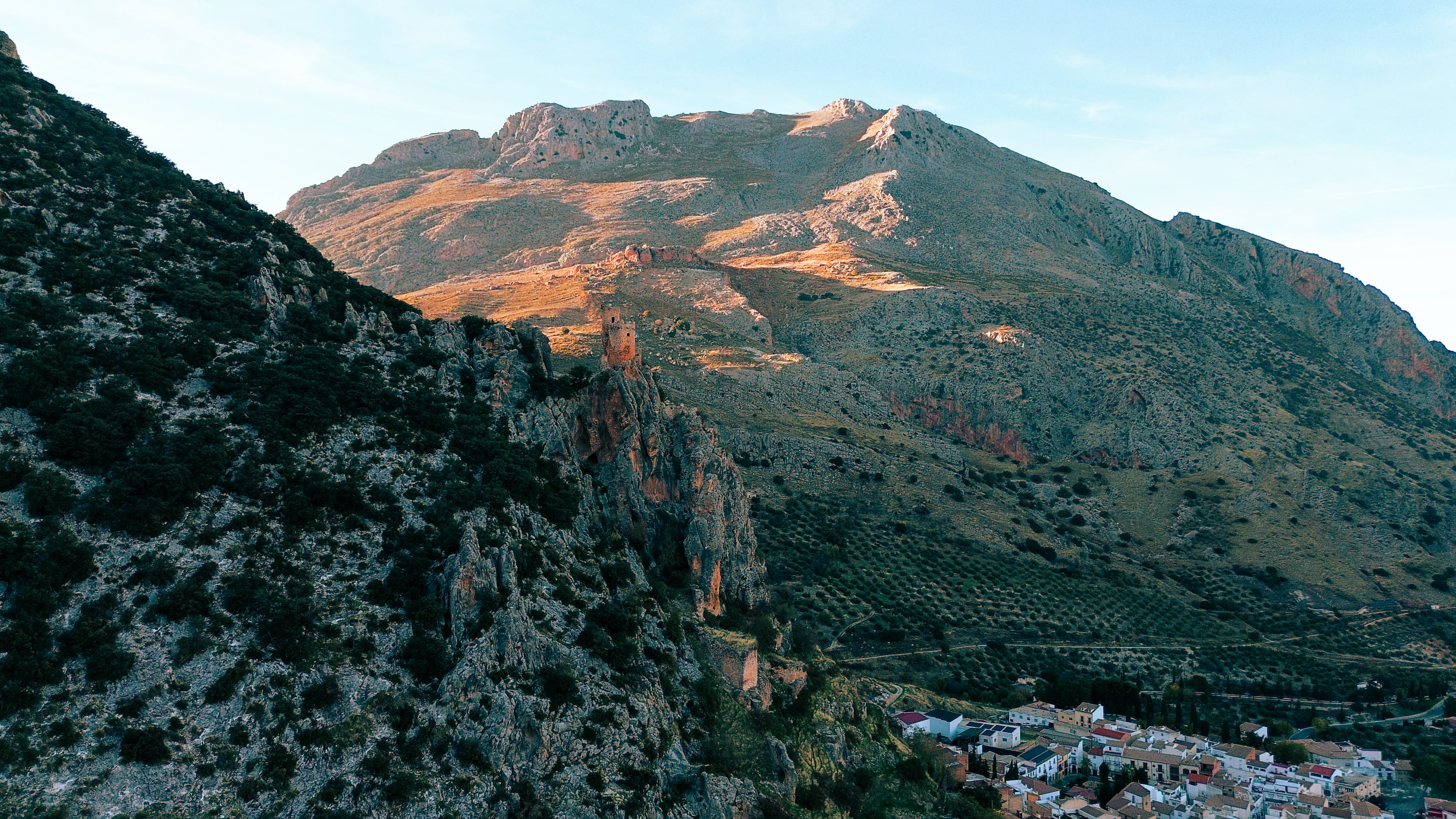 Gran montaña de laderas rocosas y vegetación dispersa, iluminada por una luz suave que resalta sus relieves. En la parte baja aparecen un escarpe y algunas construcciones del núcleo urbano, integradas en el paisaje.