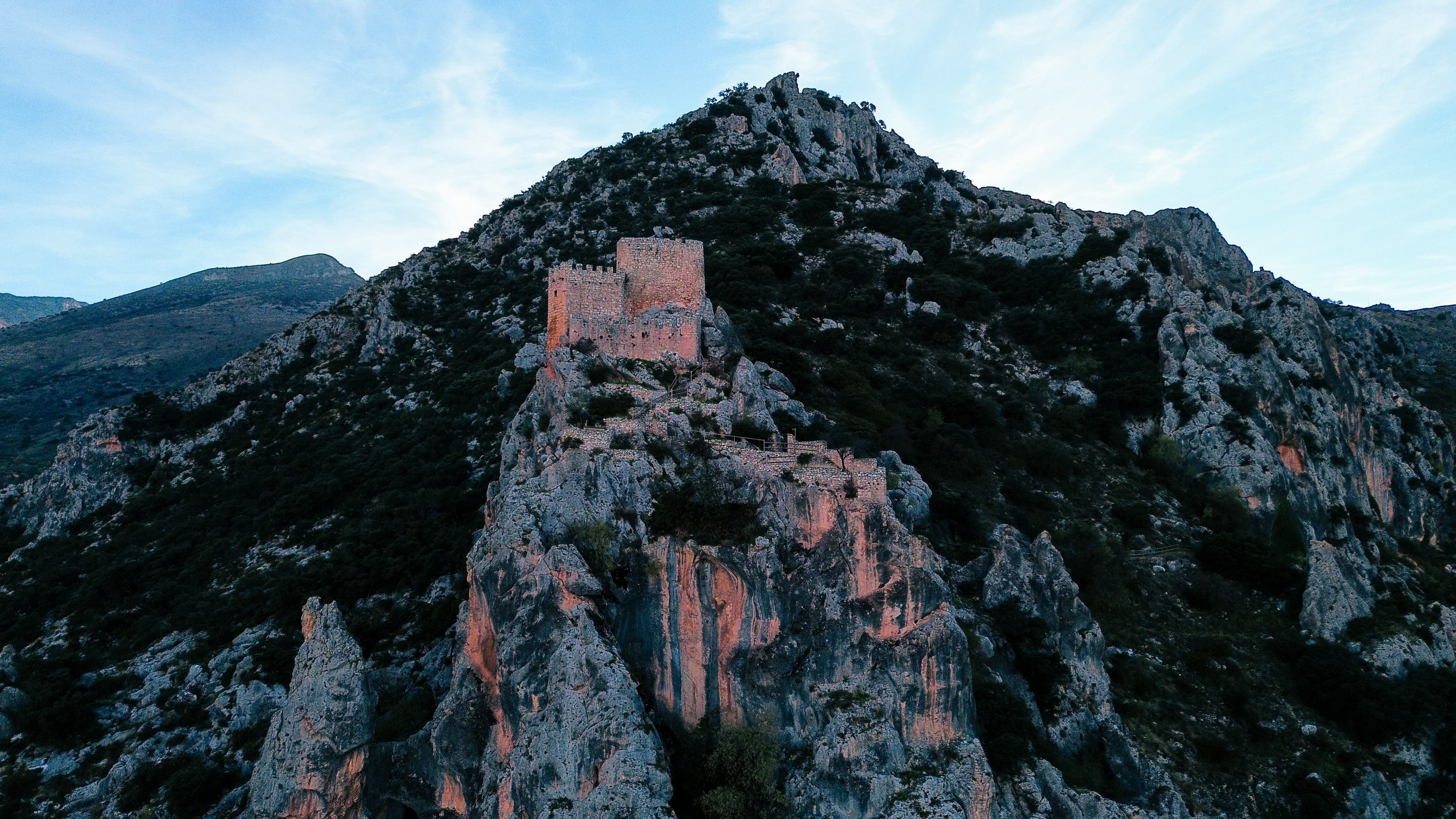 Gran montaña de laderas rocosas y vegetación dispersa, iluminada por una luz suave que resalta sus relieves. En la parte baja aparecen un escarpe y algunas construcciones del núcleo urbano, integradas en el paisaje.