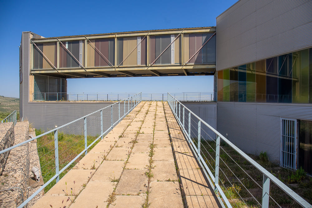 Vista exterior de un edificio moderno con fachada metálica y grandes ventanales de colores. En primer plano aparece una rampa de acceso con barandillas, rodeada por un entorno abierto y con paisaje de olivar al fondo.
