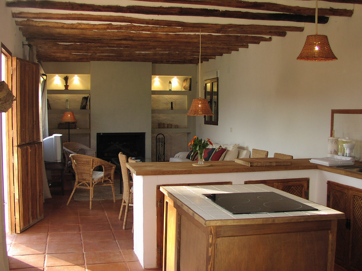 Espacio diáfano que integra cocina, comedor y zona de estar, con una isla central y acabados en madera. El techo con vigas vistas refuerza el carácter rústico del conjunto, mientras que la chimenea al fondo aporta calidez. La distribución abierta favorece la convivencia y la comodidad durante la estancia.