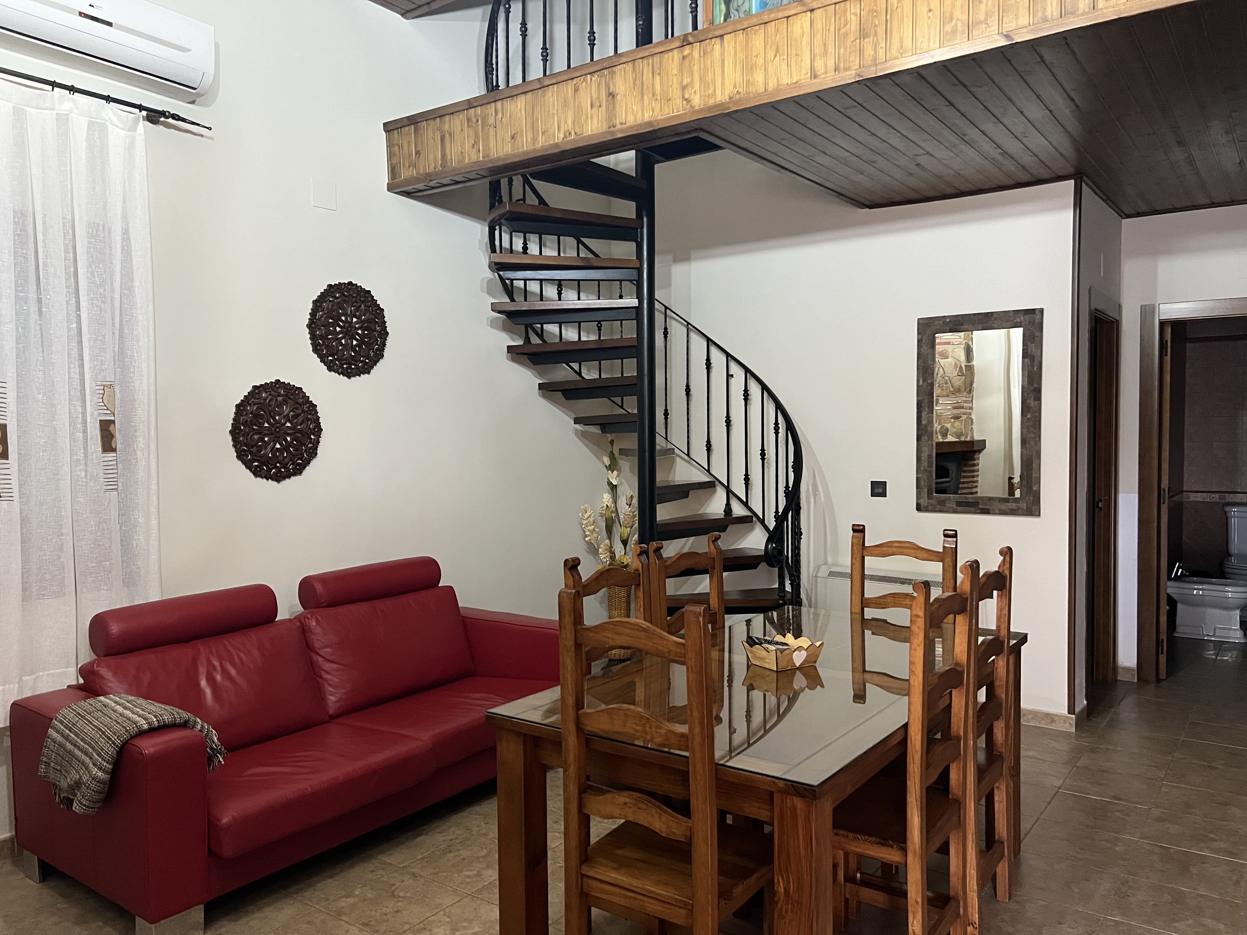 Espacio interior con mesa de comedor, sofá y una escalera de caracol que sube a un altillo, combinando estilo rústico y funcional.