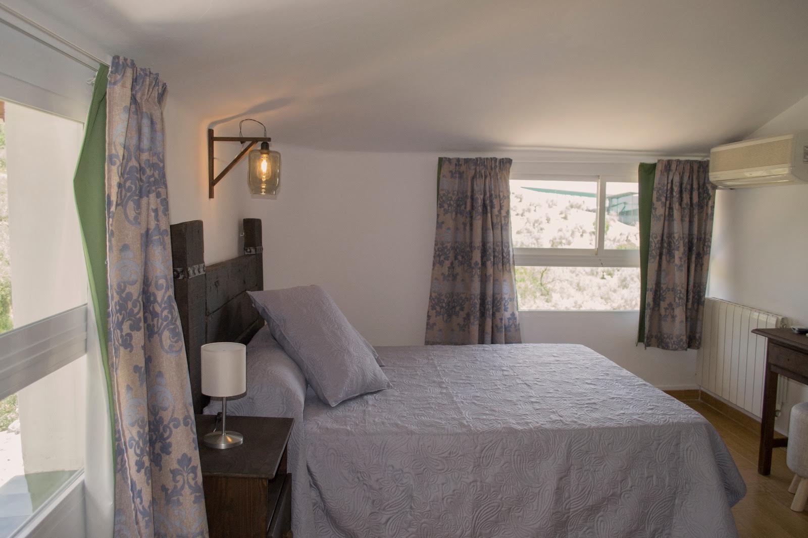 Habitación doble con mayor amplitud, equipada con cama de matrimonio, mobiliario de madera y decoración sobria. Dispone de varias ventanas que permiten la entrada de luz natural y ofrecen vistas al entorno, además de contar con elementos como aire acondicionado y escritorio. La estancia combina confort y funcionalidad en un ambiente acogedor.