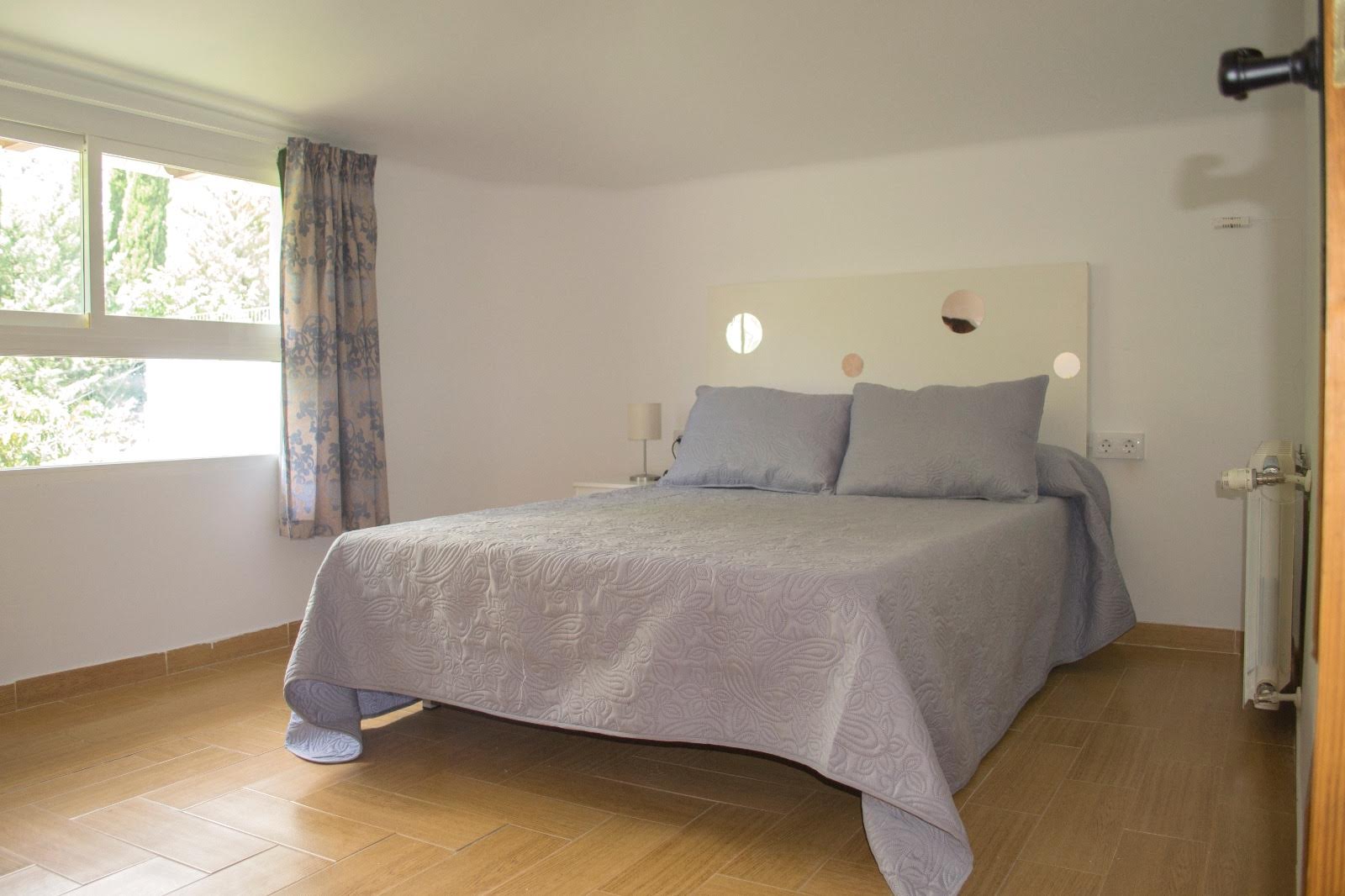 Dormitorio de estilo sencillo y luminoso, con una cama de matrimonio como elemento central, vestida con colcha en tonos neutros y acompañada de dos mesitas auxiliares con lámparas. La estancia destaca por la entrada de luz natural a través de una ventana lateral con vistas al exterior, lo que aporta sensación de amplitud y tranquilidad. Los acabados son funcionales, con suelo cerámico y una decoración minimalista orientada al descanso.
