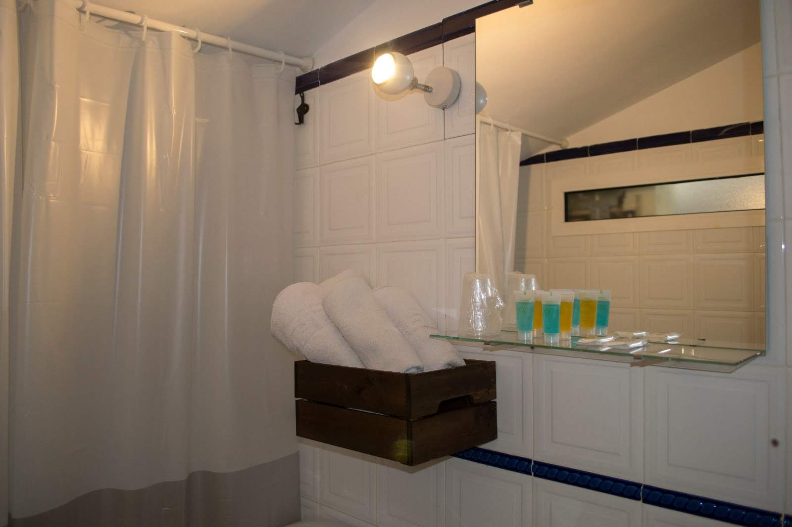 Baño completo de carácter práctico, equipado con ducha con cortina, lavabo con espejo amplio y estantería de cristal. Sobre el lavabo se disponen amenities y toallas perfectamente organizadas, lo que refuerza la sensación de limpieza y cuidado. La iluminación es cálida y el diseño responde a una estética sencilla y funcional.