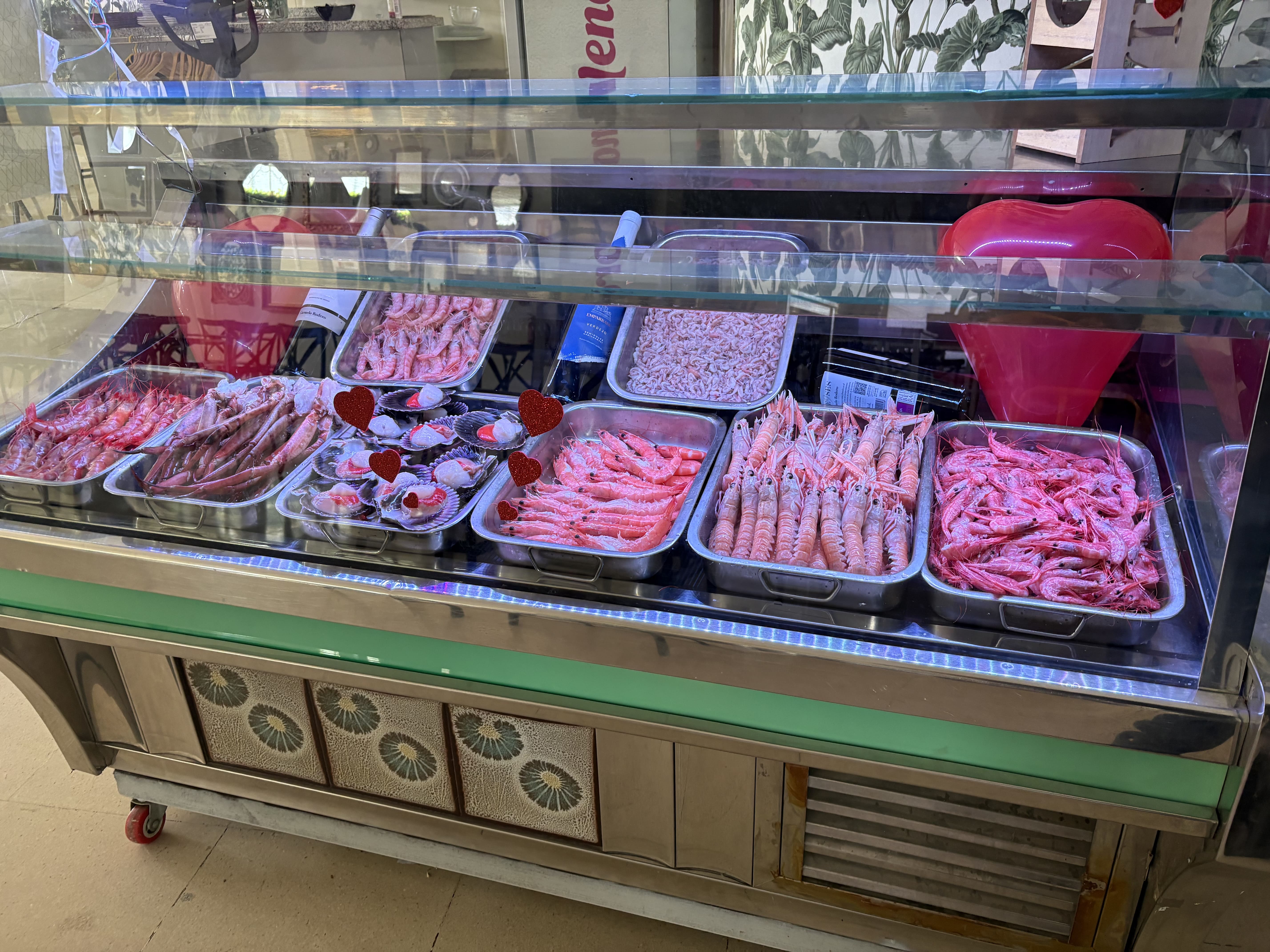 Vitrina refrigerada con bandejas de marisco (gambas, langostinos y cigalas), decorada con pequeños corazones rojos.