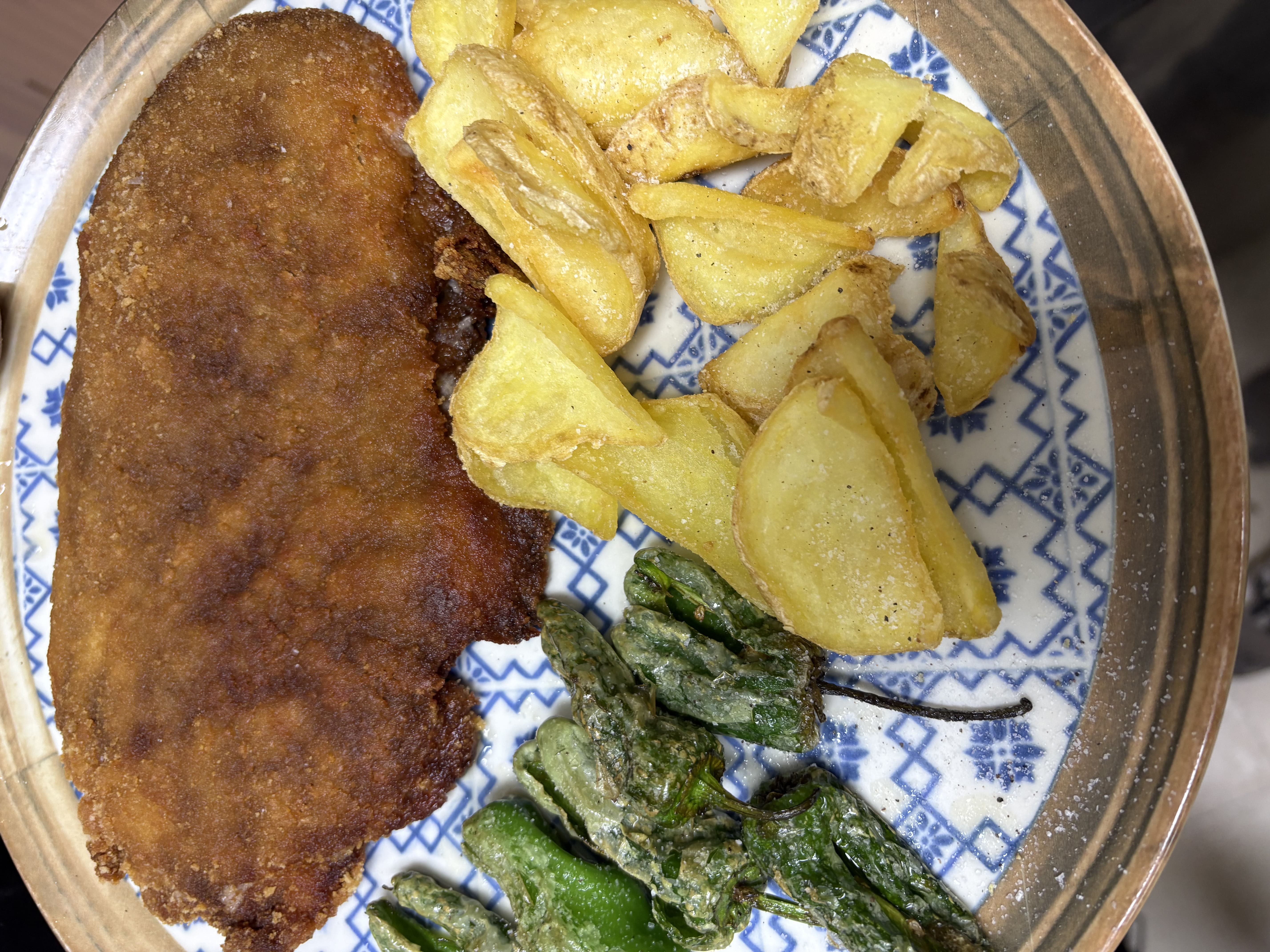 Plato con un filete empanado grande, acompañado de patatas fritas y pimientos verdes fritos.