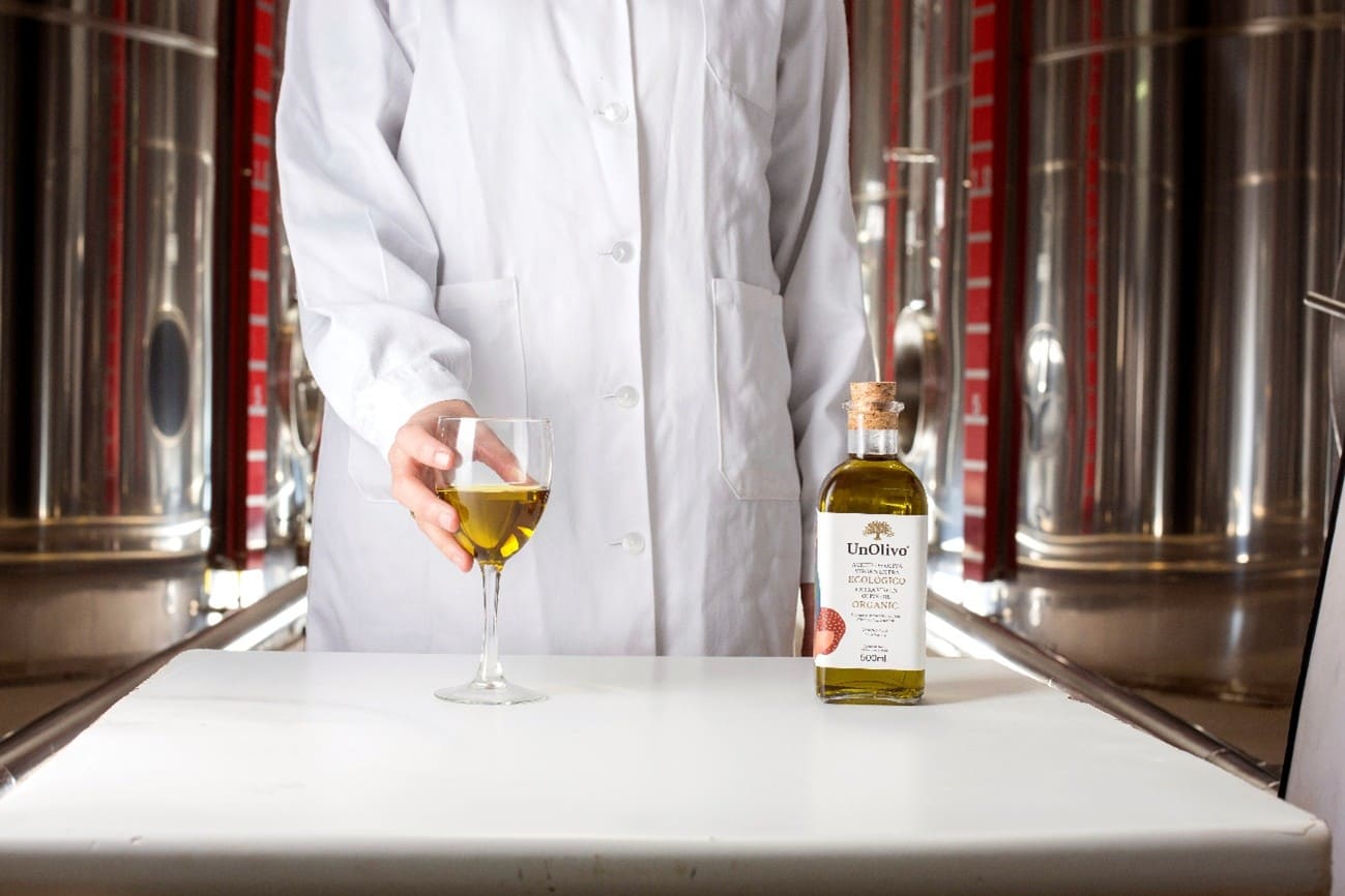 Un hombre está sosteniendo una botella de aceite de oliva virgen extra de UnOlivo en una mano y una cesta con aceitunas verdes y moradas en la otra. La escena tiene de fondo un paisaje de olivares, con un cielo claro y montañas a lo lejos, capturando la esencia del olivar.