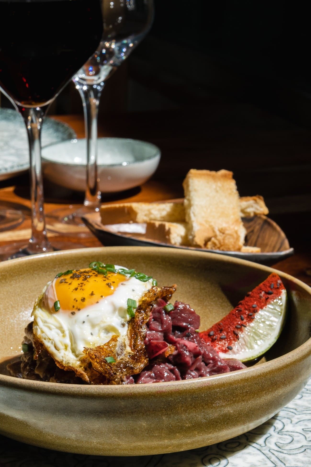 Plato gastronómico con carne, huevo frito y acompañamiento, servido junto a una copa de vino.