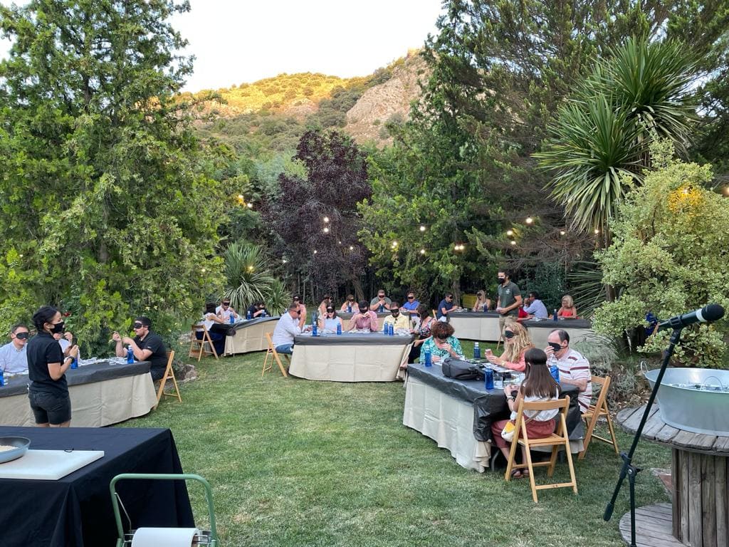 Grupo de visitantes sentados en mesas al aire libre participan en una degustación de aceite, en un entorno ajardinado con vegetación y ambiente tranquilo.