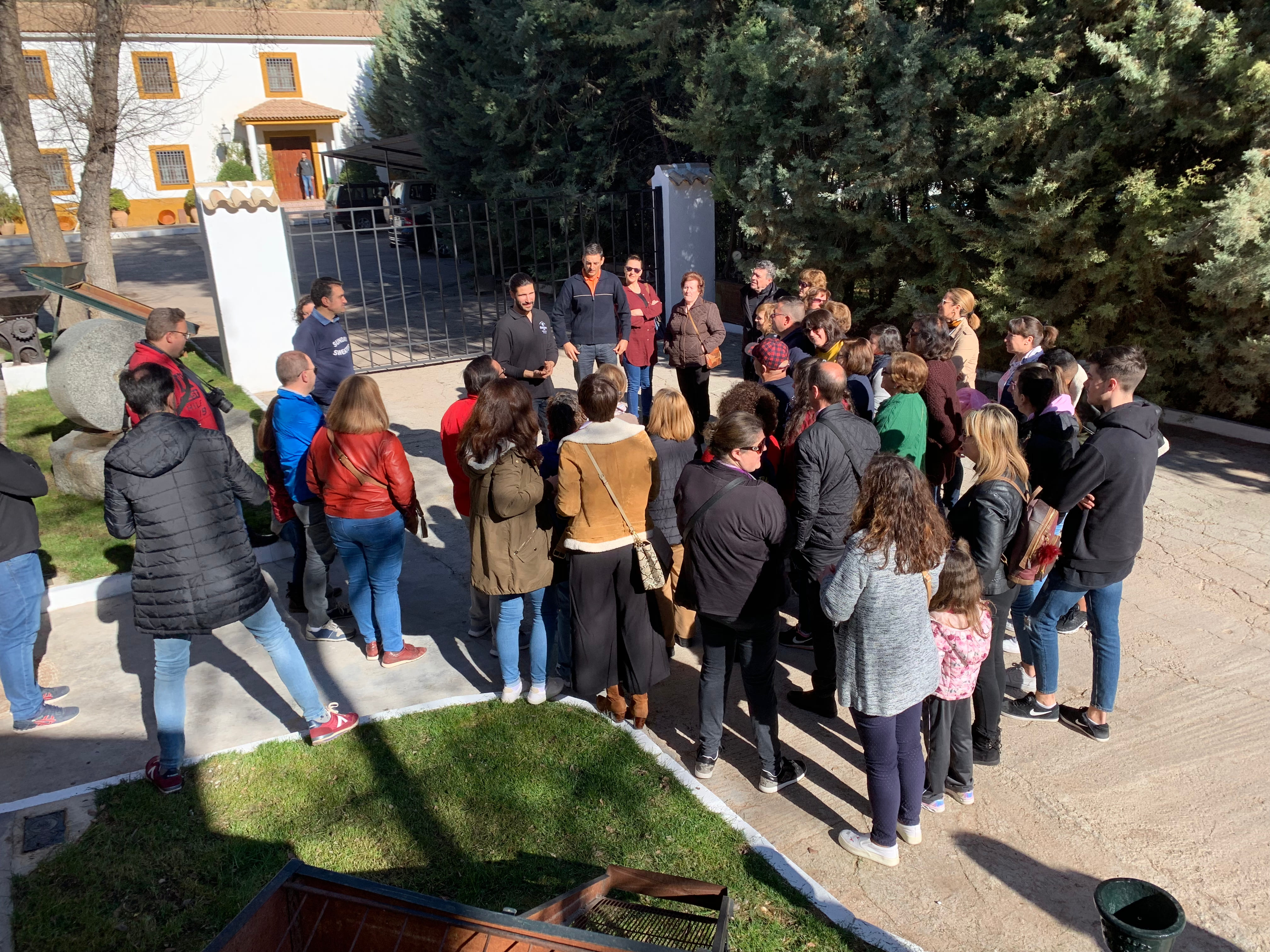 n grupo de visitantes atiende a un guía en el exterior de una almazara, durante una explicación sobre el proceso o la cultura del aceite.
