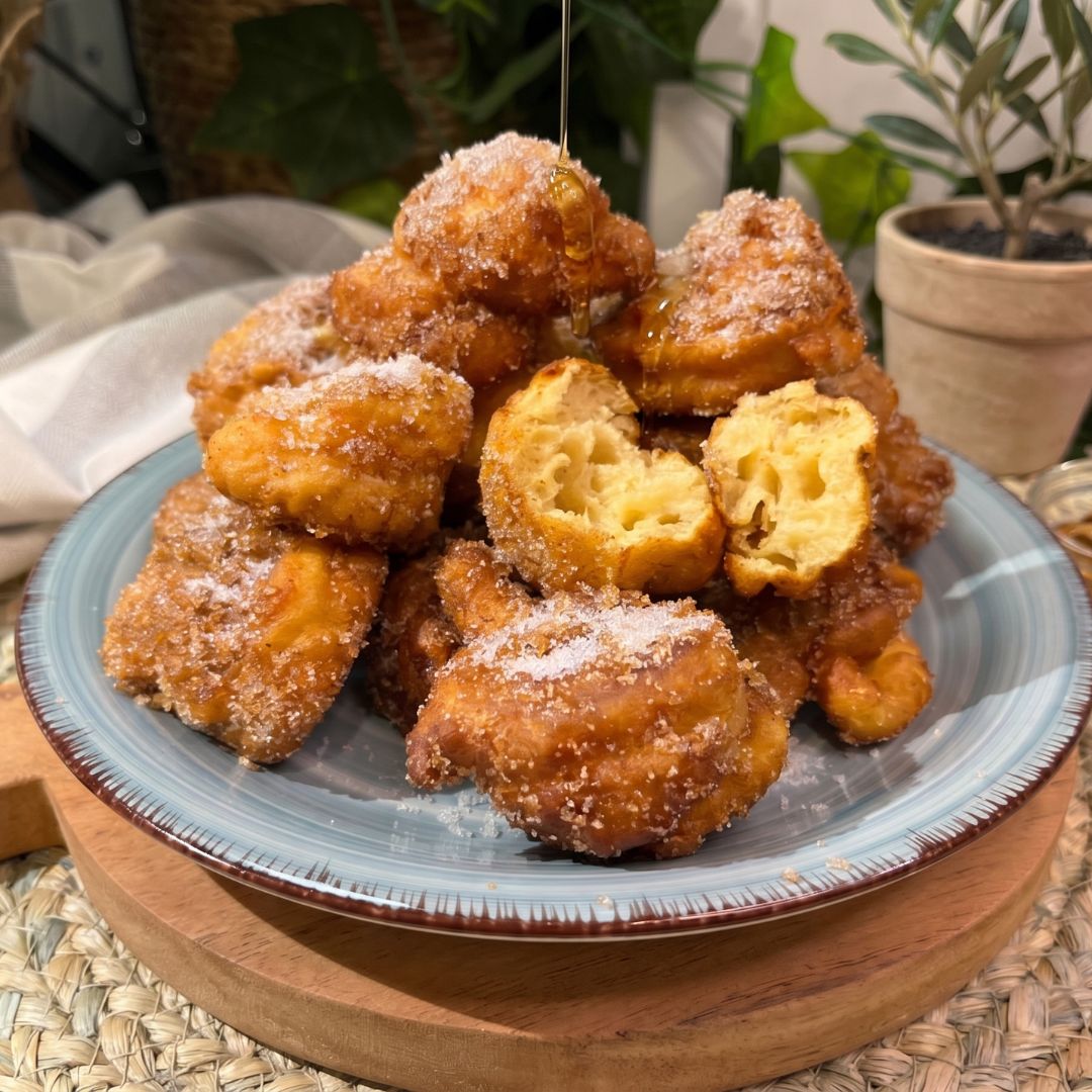 Un plato de dulce frito (tipo buñuelos o pestiños en trocitos) dorados y cubiertos de azúcar, apilados en forma de montaña sobre un plato azul. Encima cae un hilo de miel y uno de los dulces aparece abierto por la mitad, mostrando su interior esponjoso. A un lado se ve un cazo con miel y un palito mielero, con fondo de cocina rústica.