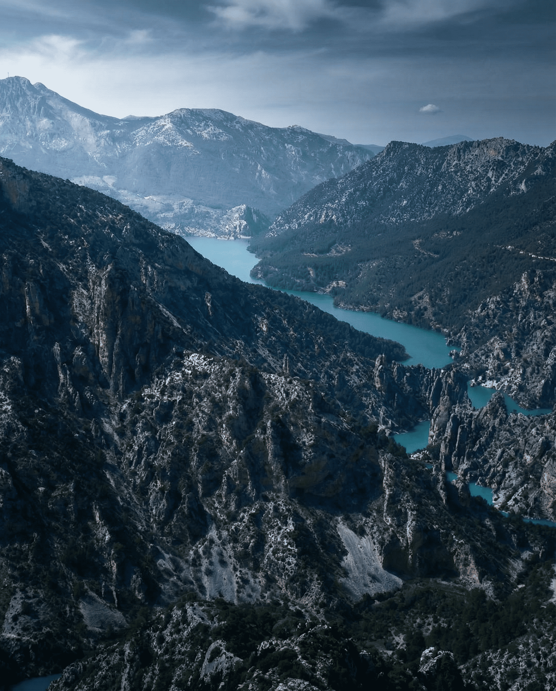 Vista panorámica de un embalse de aguas turquesas que serpentea entre montañas escarpadas y laderas rocosas. El paisaje aparece envuelto en una luz suave y un cielo cubierto, resaltando la profundidad del valle y la fuerza del relieve.