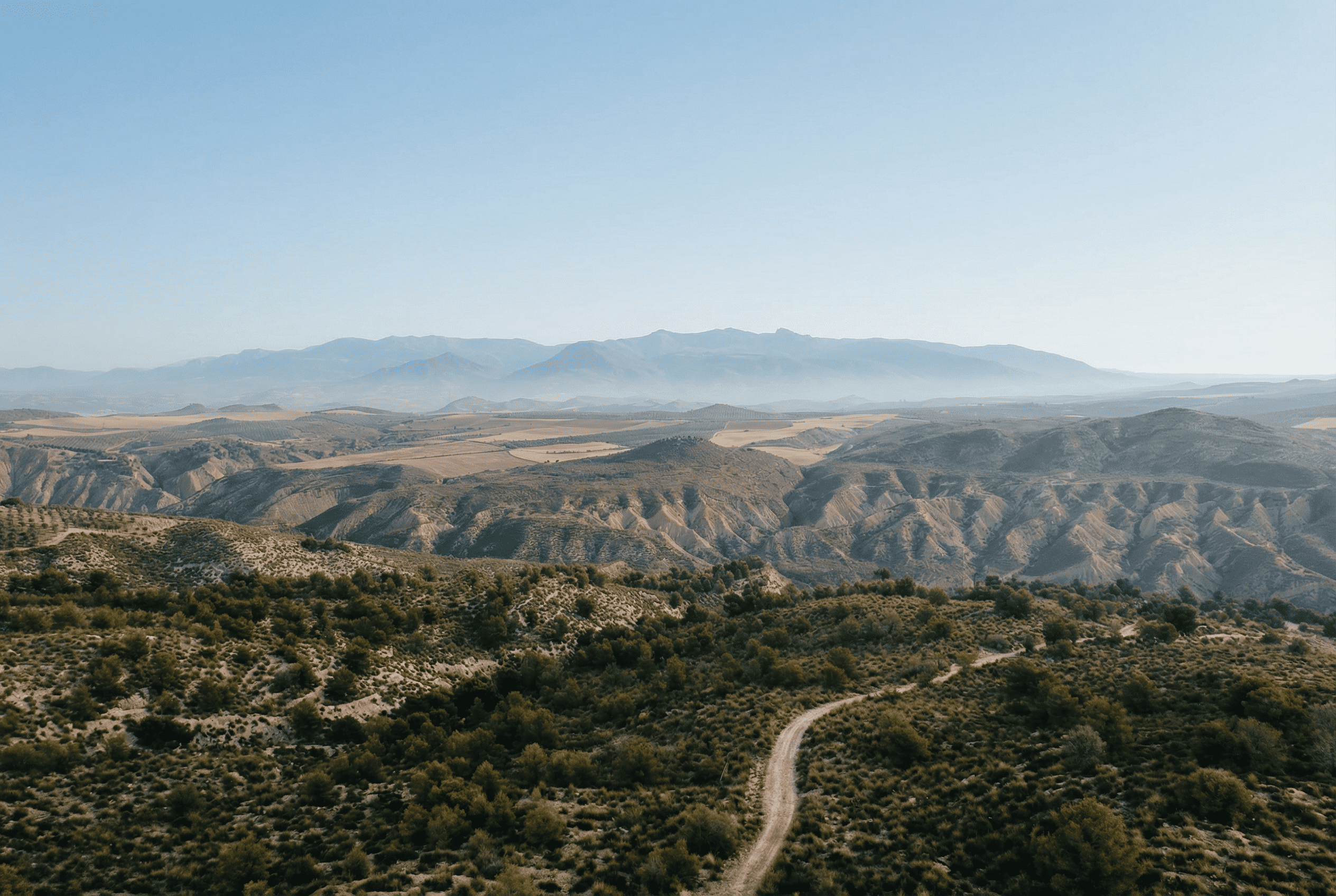 Vista panorámica de un paisaje abierto con colinas, barrancos y caminos que se extienden hasta el horizonte. Al fondo se distinguen varias sierras difuminadas por una ligera bruma bajo un cielo despejado.