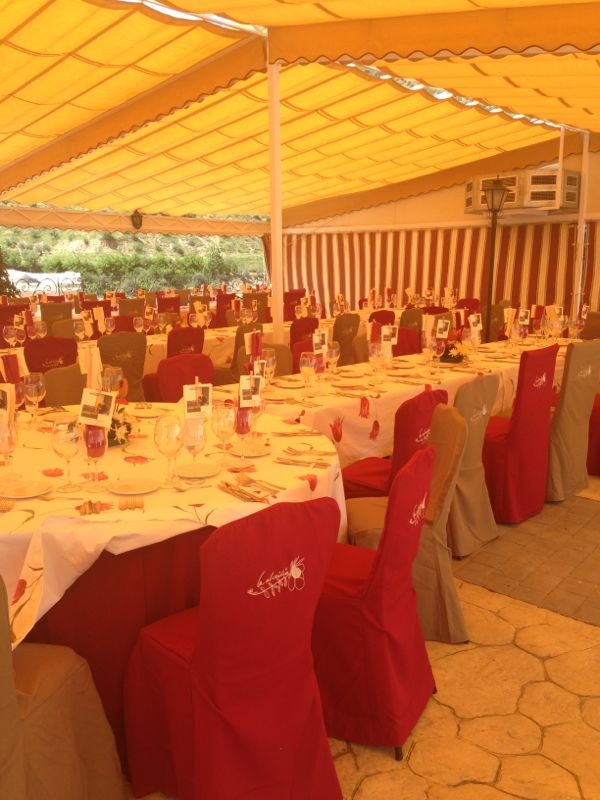 Terraza de restaurante preparada para una celebración, con mesas redondas vestidas, vajilla colocada y sillas cubiertas en tonos rojos y beige bajo un toldo amarillo.