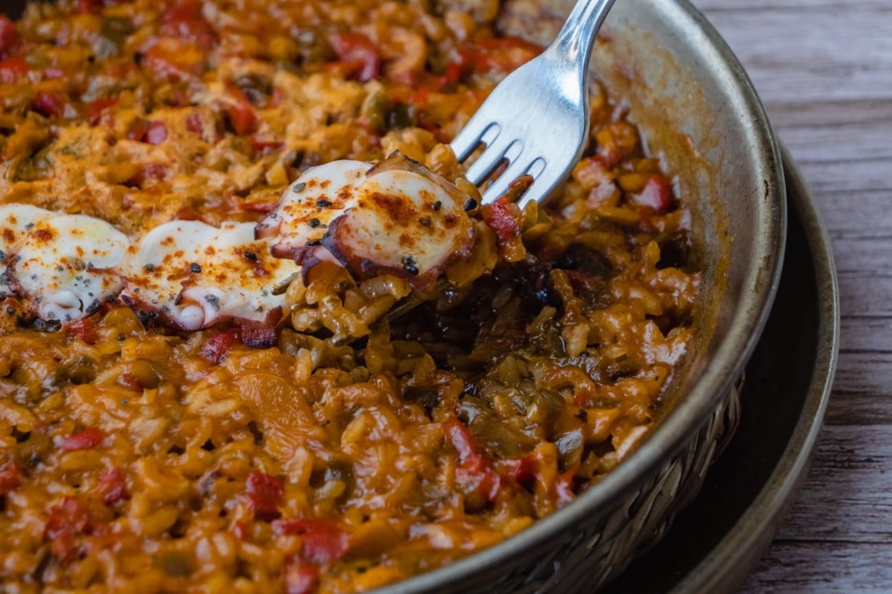 Primer plano de una sartén con un arroz meloso de tonos rojizos y verduras; un tenedor levanta un bocado con un trocito de pulpo, con el resto del plato de fondo sobre una mesa de madera.