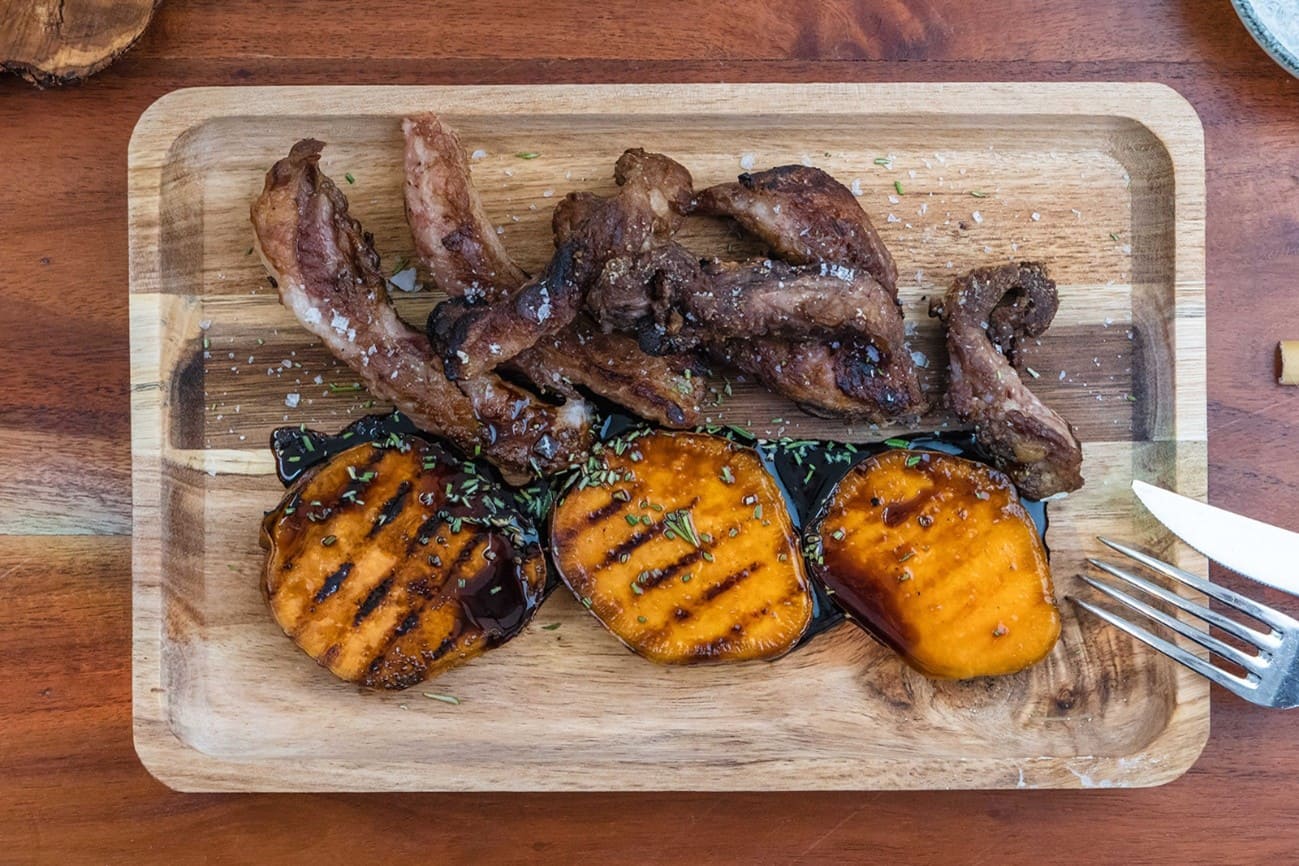 Tabla de madera con carne asada en tiras (tipo costilla o secreto) y tres rodajas de boniato a la plancha con marcas de parrilla, acompañadas de una salsa oscura y hierbas espolvoreadas; a la derecha se ve un tenedor.