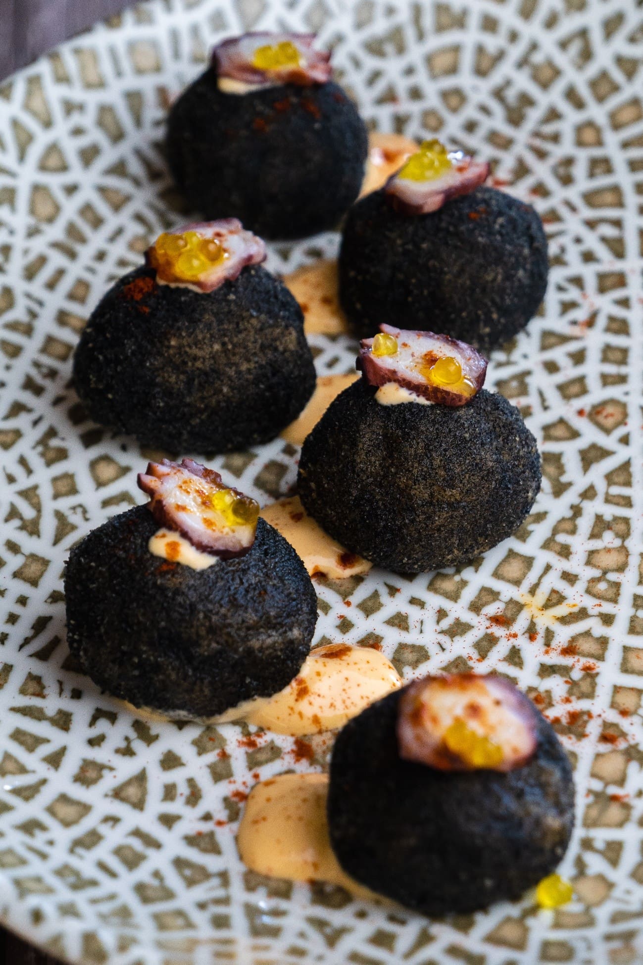 Primer plano de varias croquetas negras, redondas, servidas en una bandeja con dibujo geométrico. Van coronadas con una lámina de pulpo y pequeñas