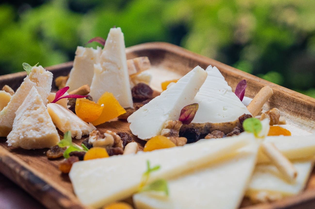 Tabla de quesos presentada sobre una base de madera, con varios cortes y cuñas de quesos blancos/curados. Se acompaña de frutos secos y pasas, dados de fruta naranja.
