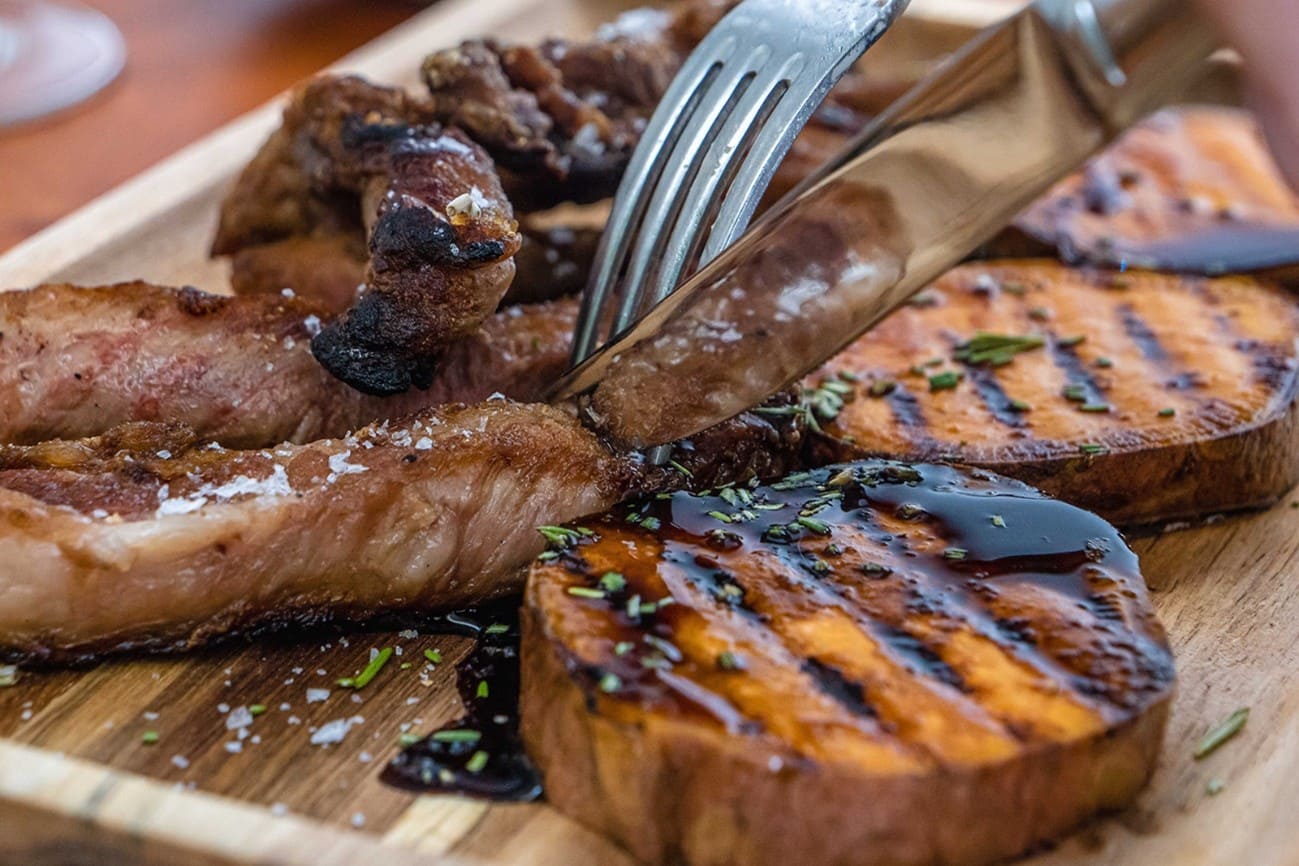 Primer plano de una tabla de madera con tiras de carne a la brasa y rodajas de boniato asado marcadas a la parrilla, bañadas con una salsa oscura y espolvoreadas con hierbas y sal en escamas.