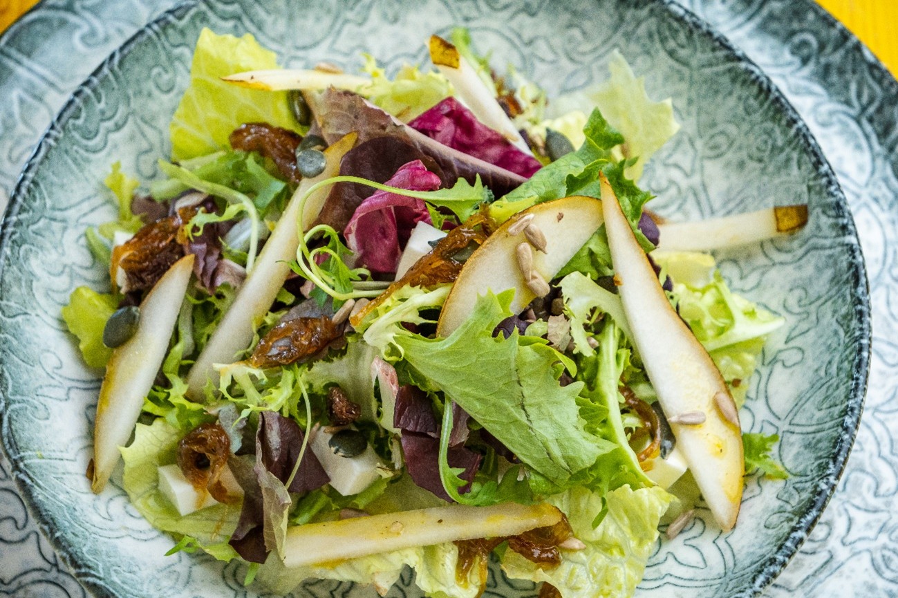 Ensalada servida en un cuenco de cerámica gris, con mezcla de hojas verdes y moradas, láminas de pera, dados de queso blanco, semillas (pipas) y trocitos oscuros tipo fruta deshidratada, todo con un aliño brillante.
