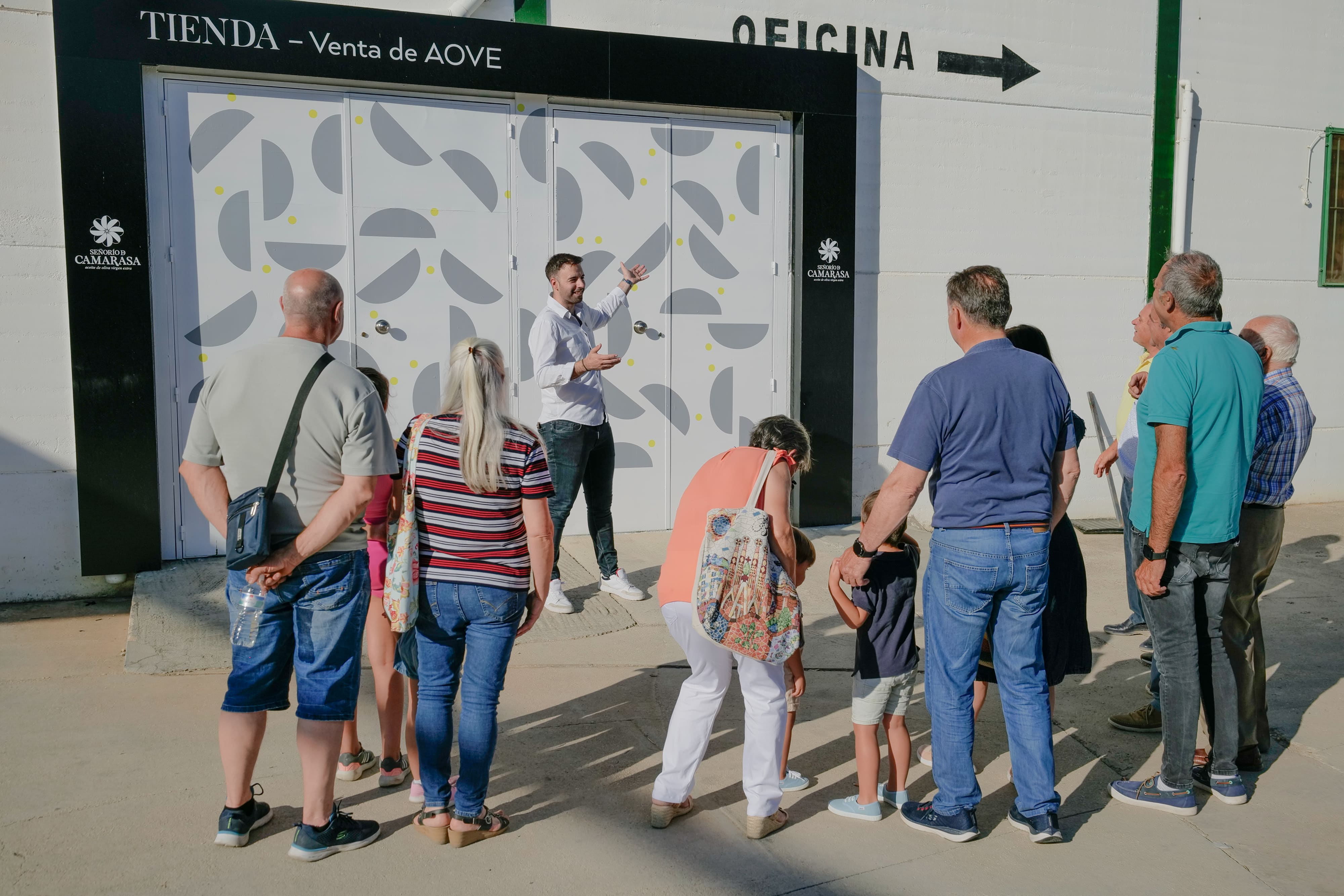 Un pequeño grupo de personas, incluyendo niños y adultos, está de pie frente a un edificio con un cartel que dice "TIENDA – Venta de AOVE". Un guía está de pie frente a ellos, hablando y señalando hacia la entrada del lugar.