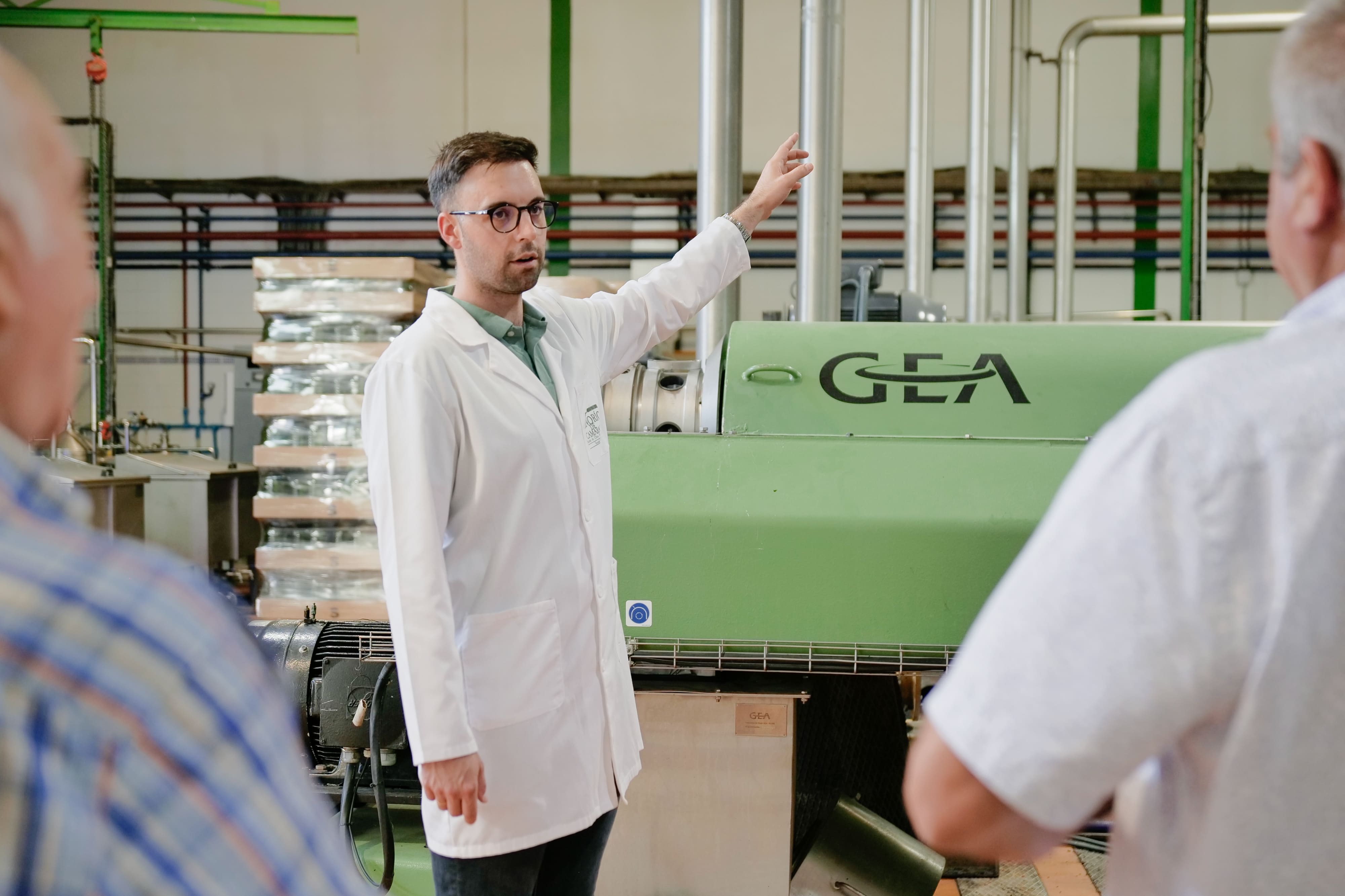 Un hombre con bata blanca está dando una explicación sobre la maquinaria de la fábrica. Está señalando una gran máquina industrial verde, mientras algunos visitantes escuchan atentamente.