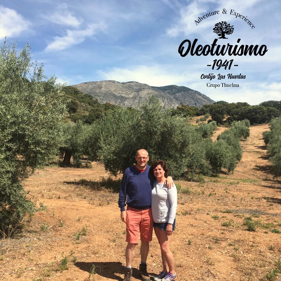 Pareja posando entre olivares con montañas al fondo, en un paisaje abierto y soleado.